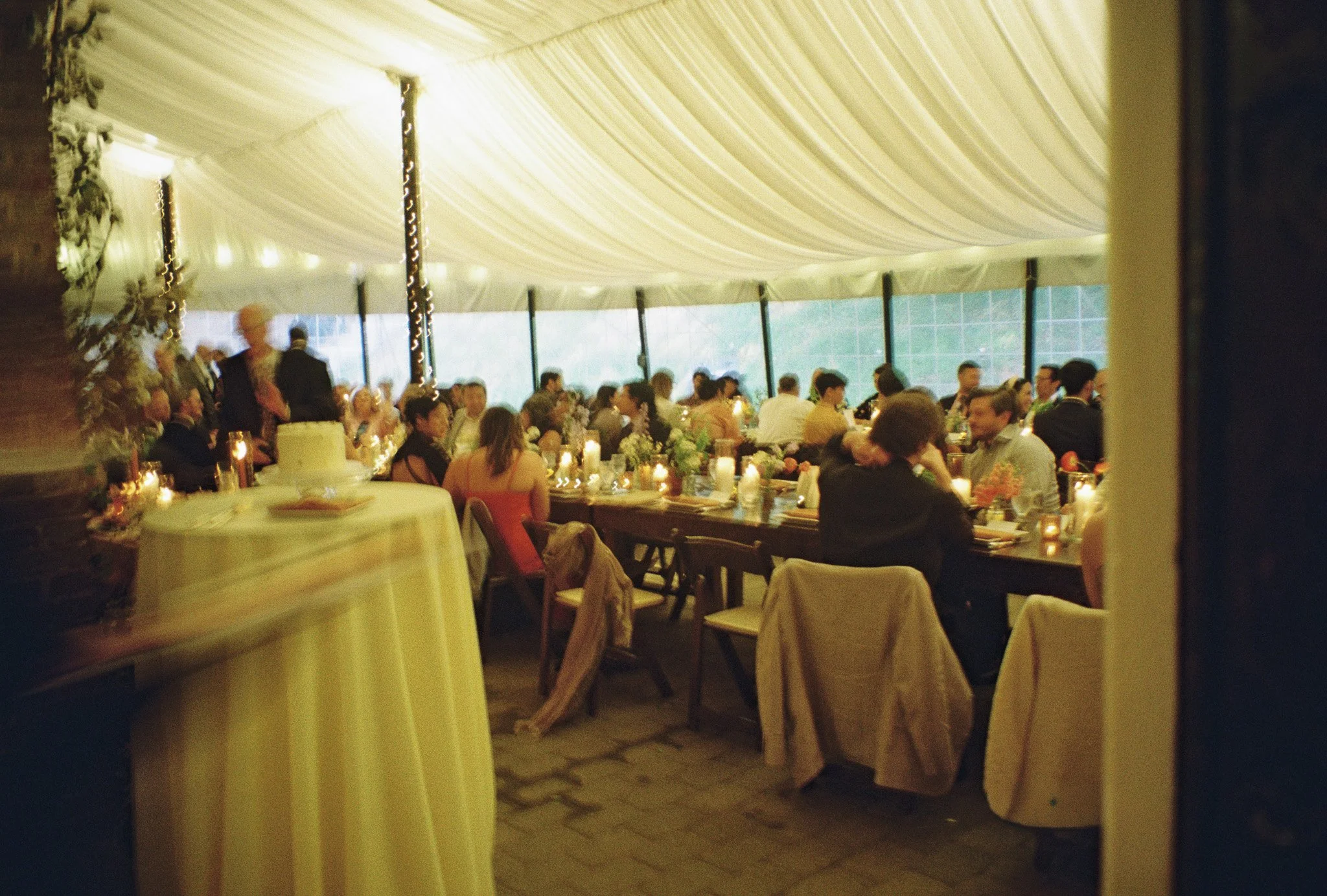 Indoor wedding reception with guests seated at tables, decorated with candles and flowers, under a tented ceiling with string lights.