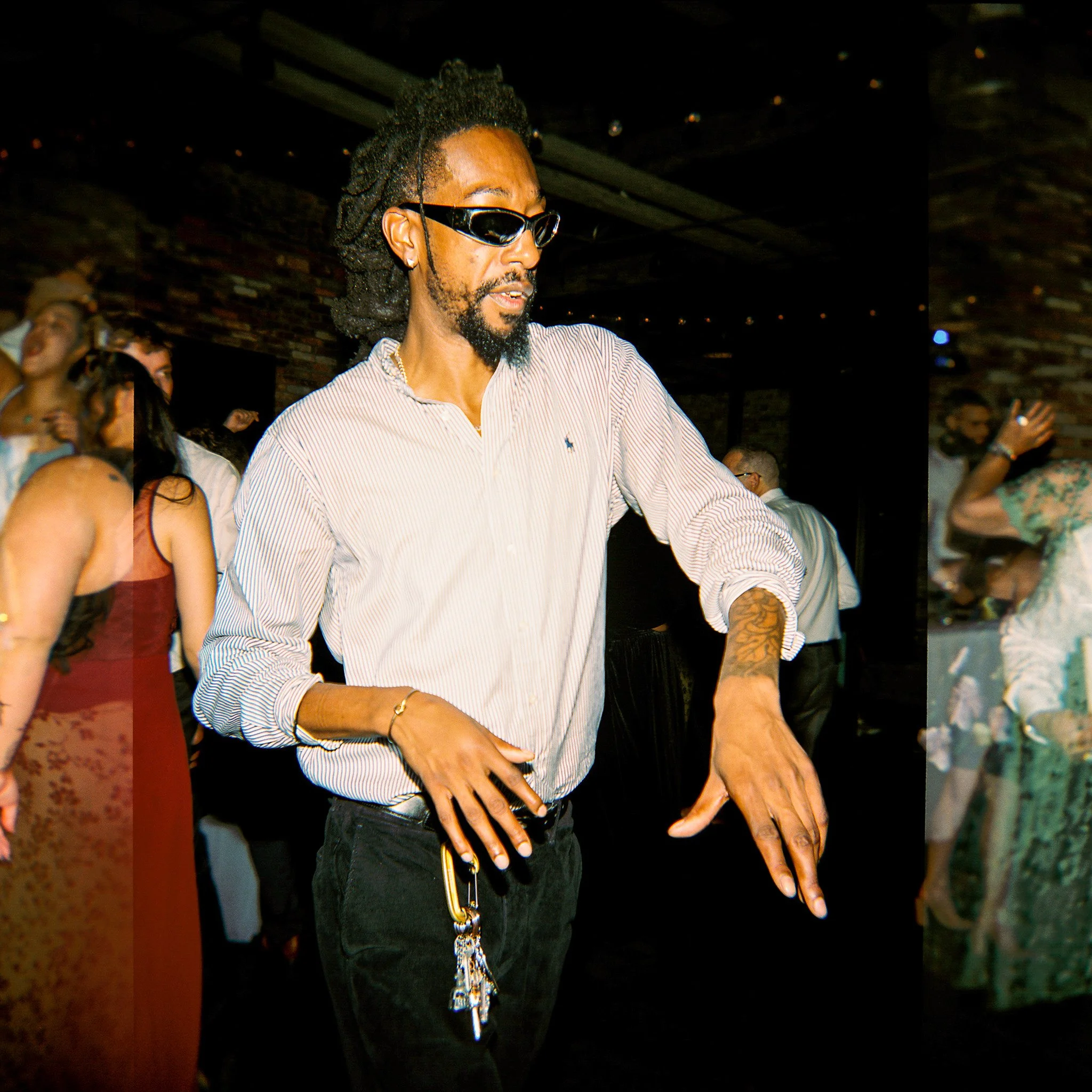 A man with dreadlocks, wearing sunglasses and a striped shirt, dancing or enjoying music at a crowded night event.