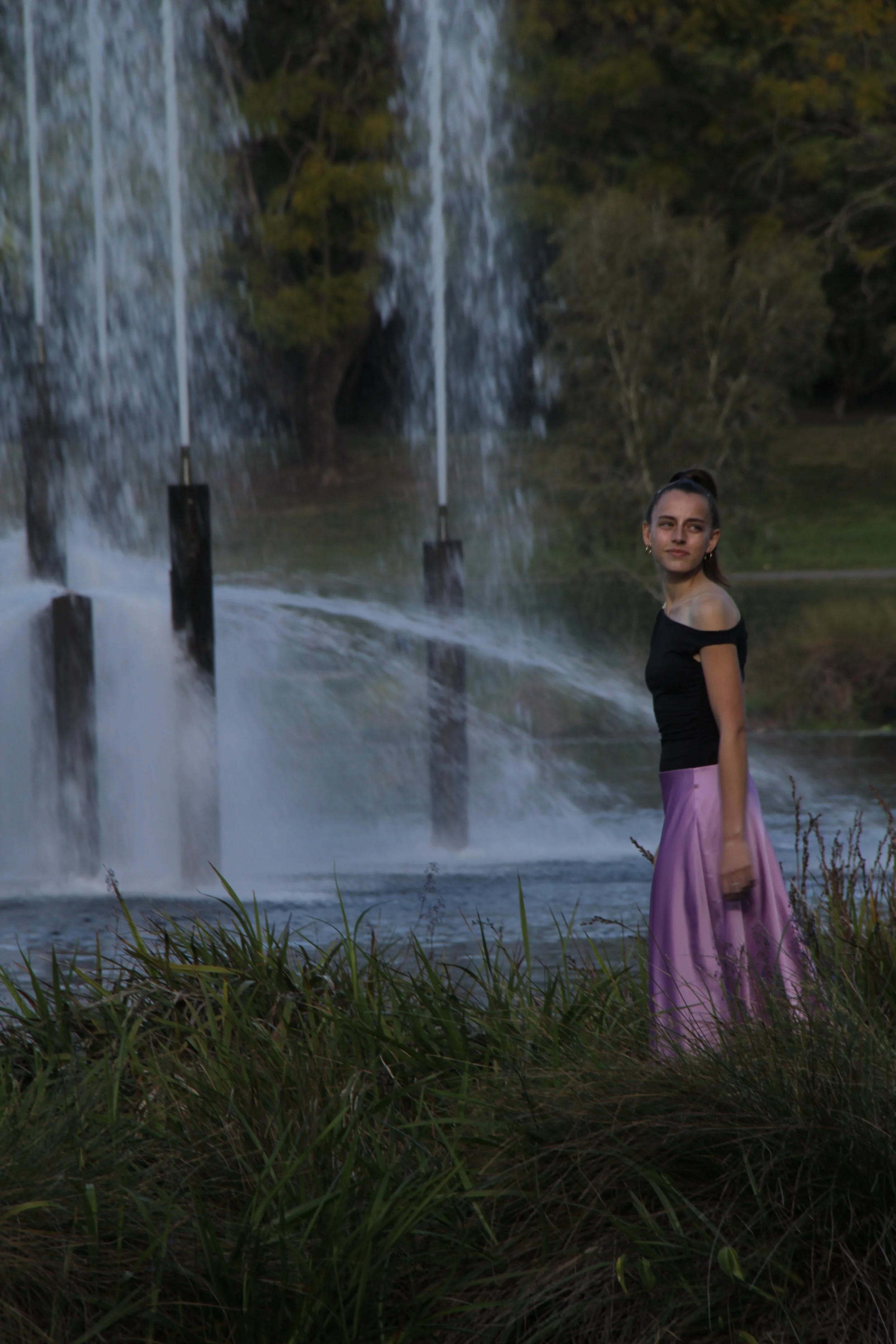 A woman standing by a waterfall surrounded by greenery, wearing a black top and purple dress, with water mist rising around her.