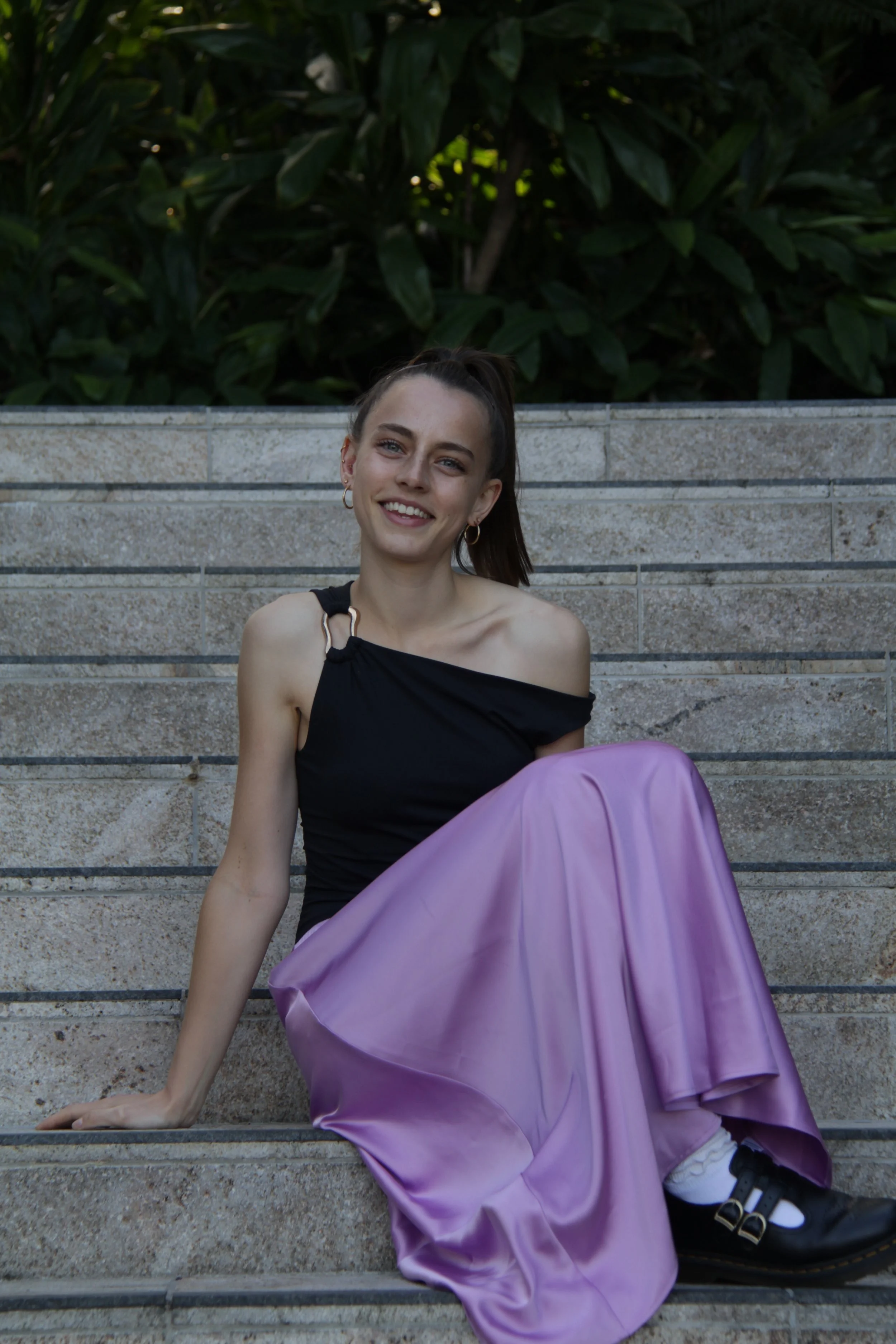 A smiling young woman with brown hair tied back, wearing a black off-shoulder top, sitting on outdoor stone steps with a purple satin skirt, and plants in the background.