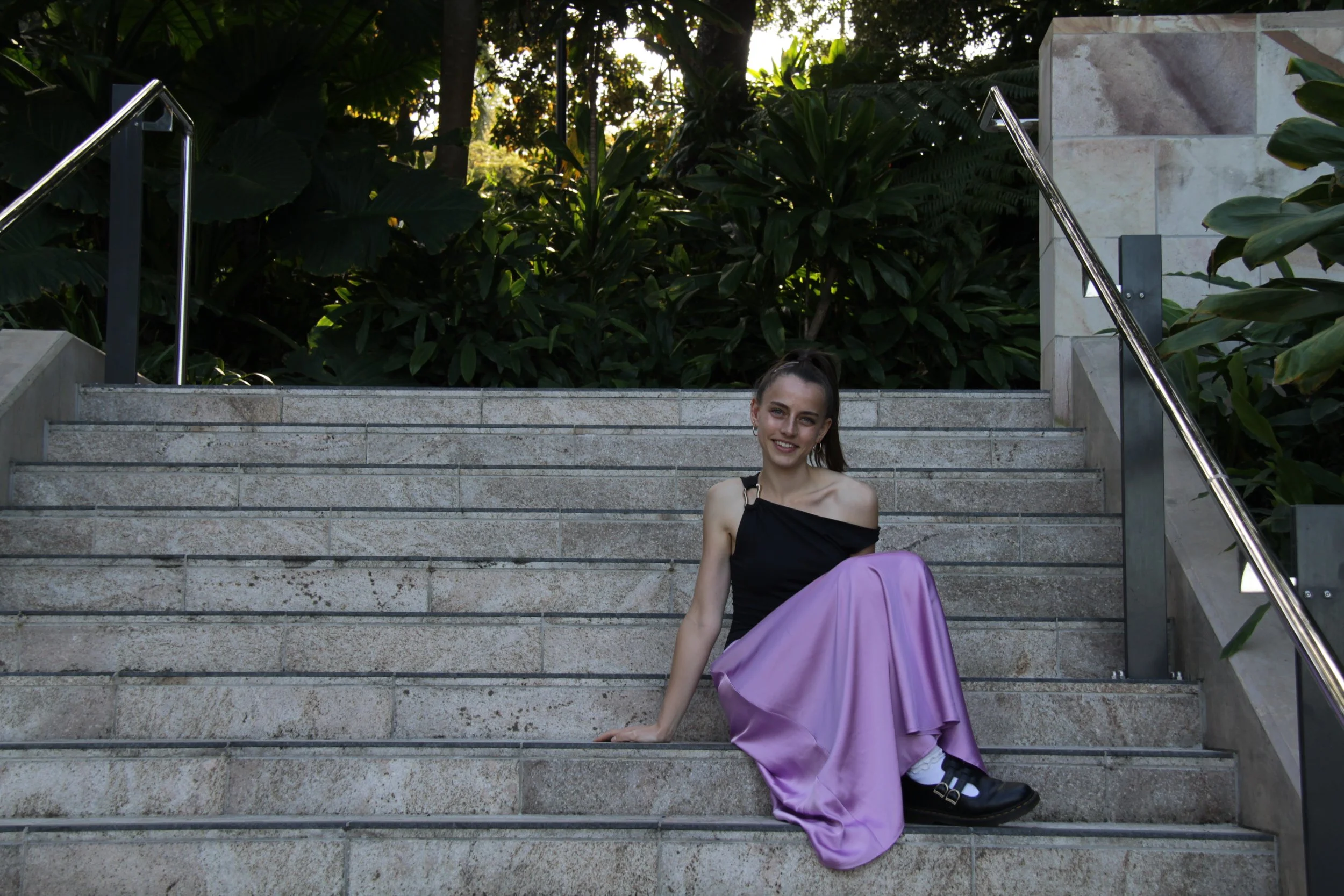 A young woman sitting on outdoor stone steps, smiling, wearing a black and purple dress, with greenery and trees in the background.