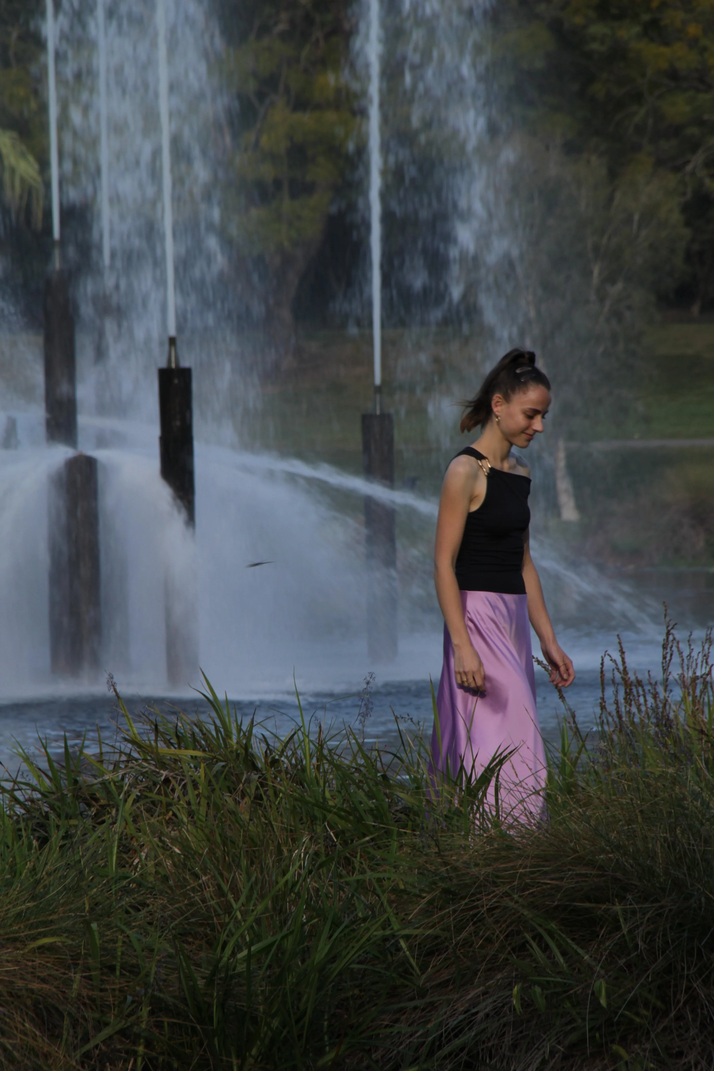 A woman in a black top and purple skirt walking on a grassy area near a waterfall with water cascading down rocks behind her.
