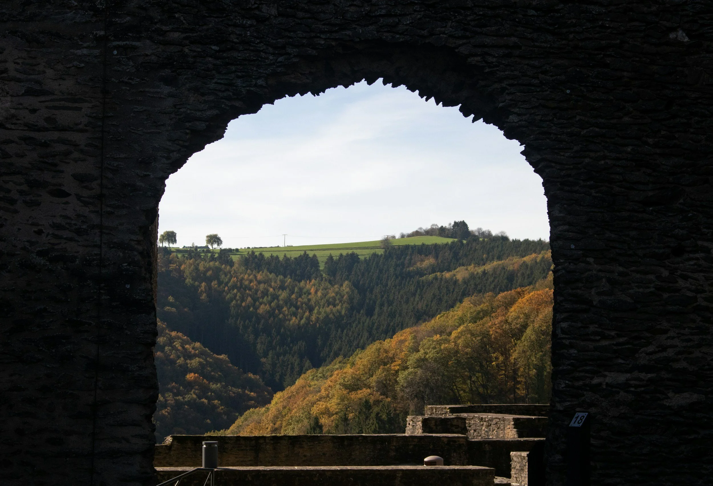 View through an arched stone window showing trees and green rolling hills in the distance under a partly cloudy sky.
