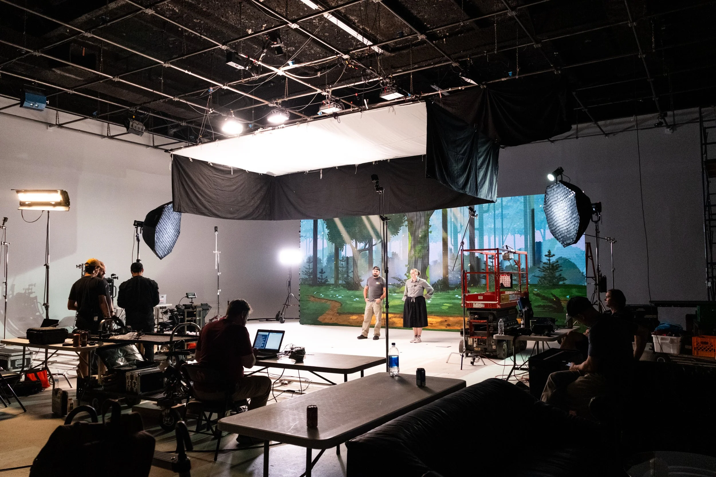 Film crew recording two actors on a soundstage with studio lights, cameras, and a forest backdrop