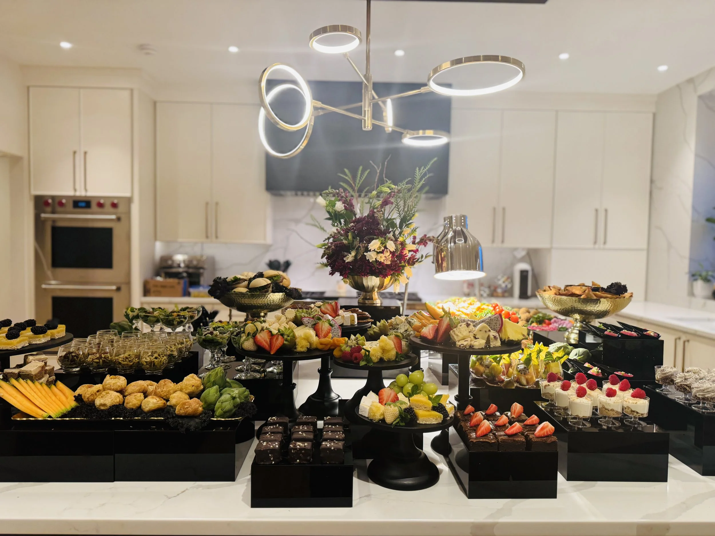 A dessert table with a variety of desserts including cupcakes, fruit, cookies, chocolates, and pastries, displayed on black and tiered stands in a modern kitchen with white cabinets and a gold and ring chandelier.