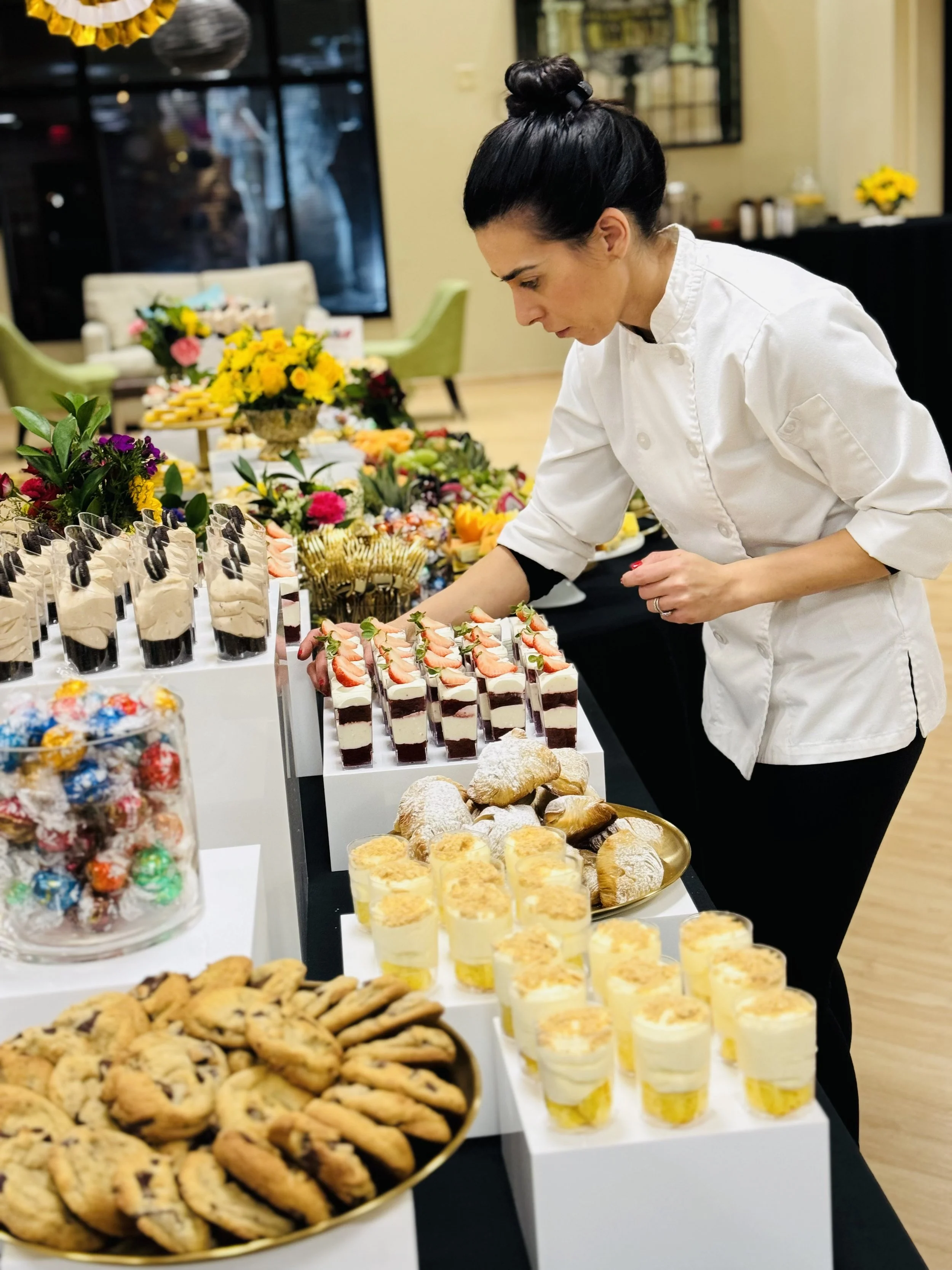 A woman in a white chef's coat arranging desserts on a table with an assortment of cookies, cakes, and pastries, with floral arrangements in the background.