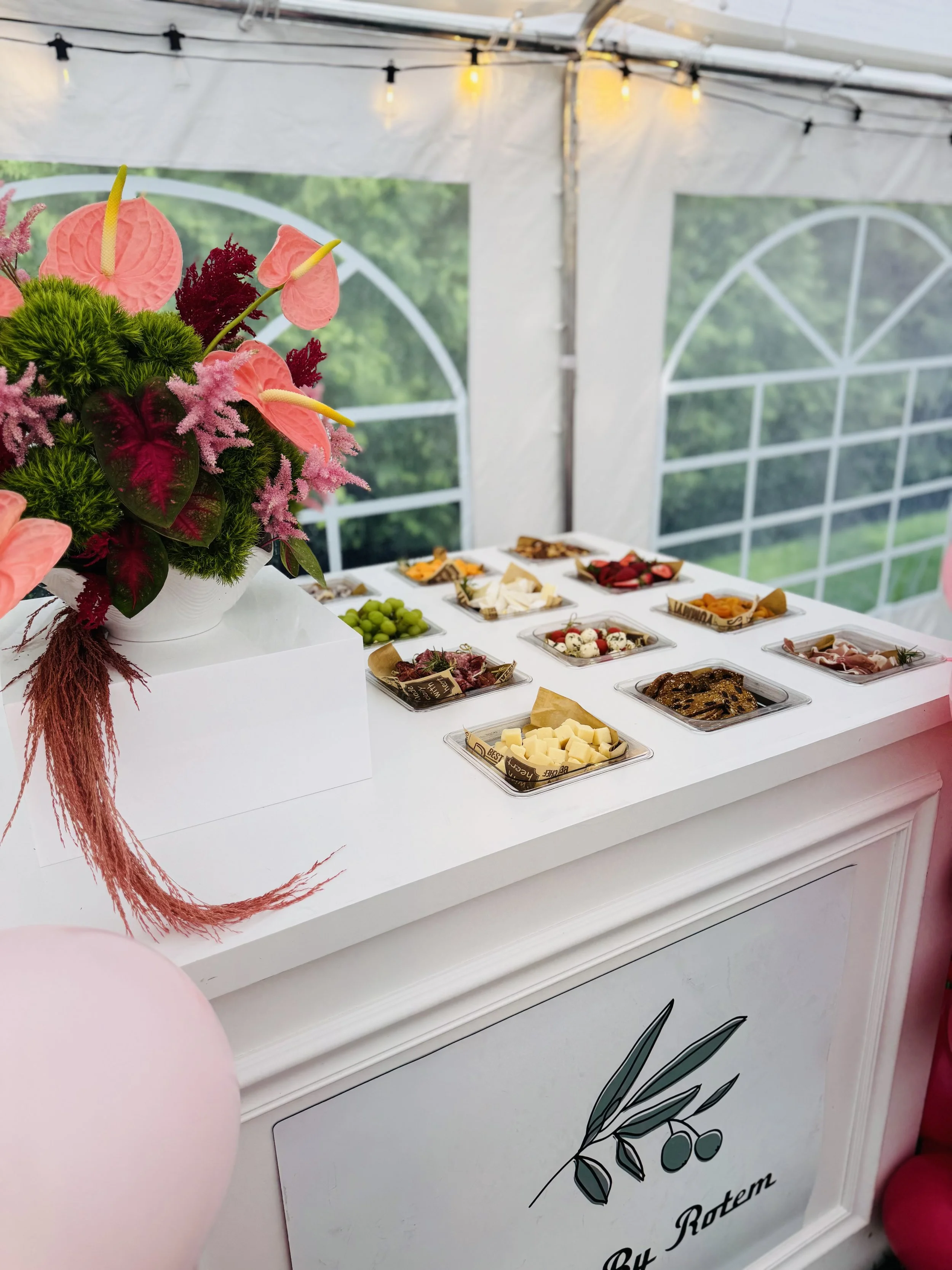 A table with a floral arrangement of pink anthuriums, pink astilbe, and green foliage. The table also has small dishes filled with various foods such as cheese, meats, and fruits. The setup is inside a white tent with arched windows and string lights overhead.