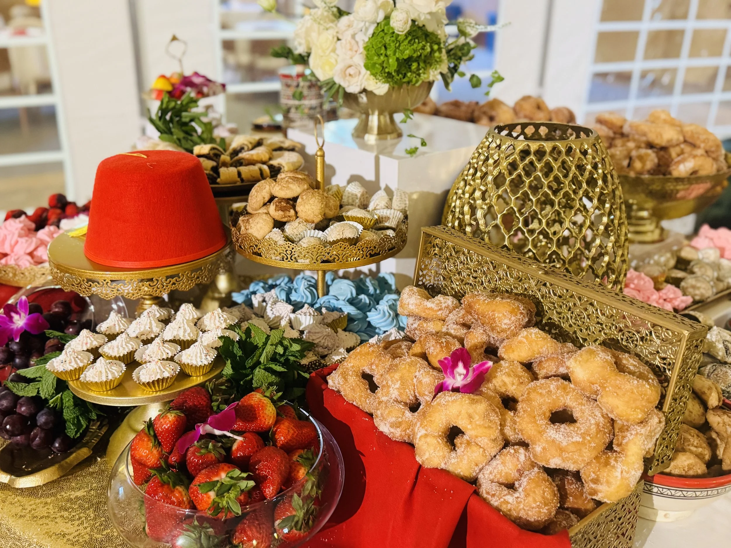 A dessert table decorated with gold ornate trays and bowls filled with various sweet treats, fresh strawberries, grapes, and flowers, with a centerpiece of white roses and green hydrangeas.
