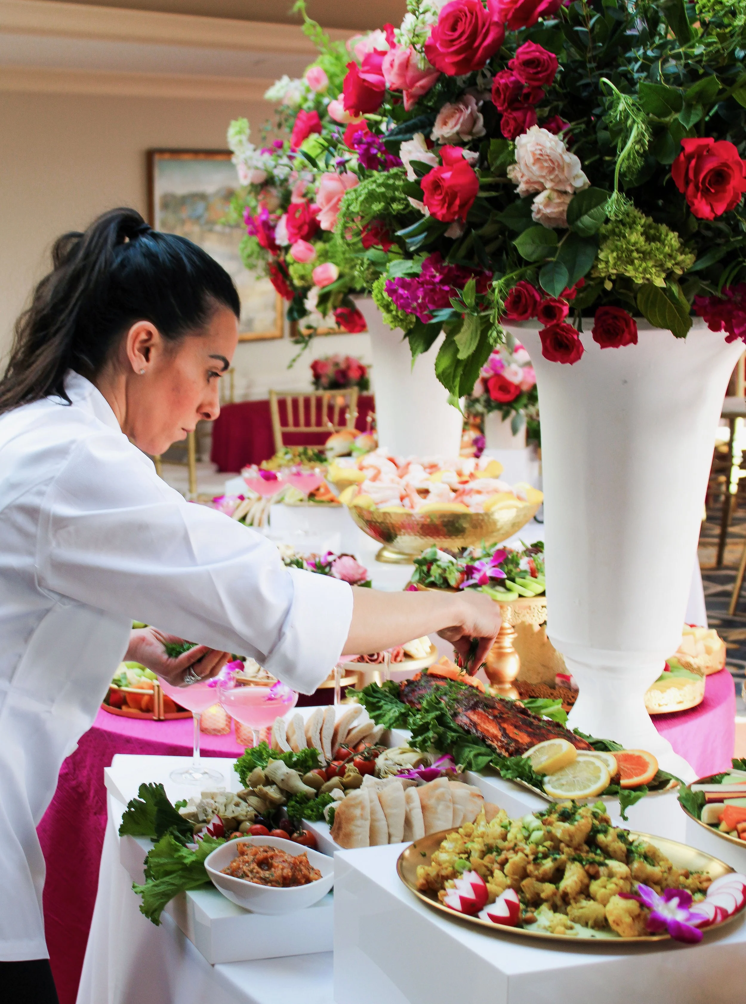 A woman in a white chef's coat is serving herself food from an elegant buffet table filled with a variety of dishes, garnished with fresh herbs, fruits, and flowers, with large white vases of vibrant pink and red flowers as decorations.