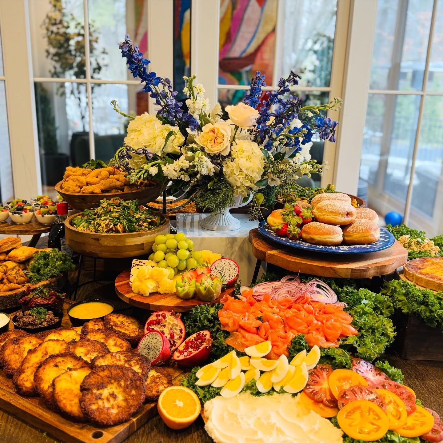A buffet table with various dishes, including a large colorful flower arrangement in a vase, a plate of mixed berries and fruit, a salad with greens and vegetables, a dish of scrambled eggs garnished with green vegetables, and other assorted foods in bowls and platters.