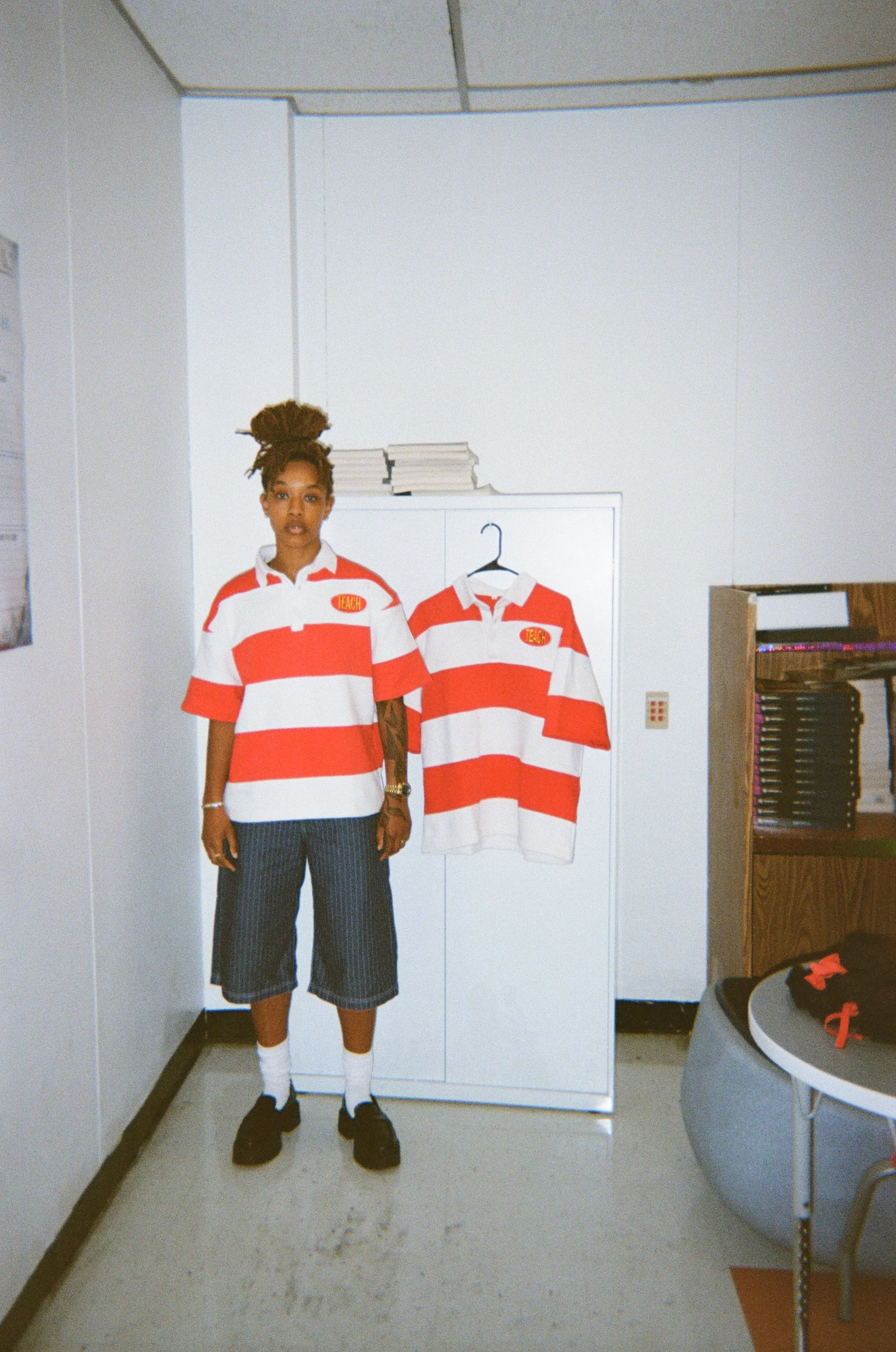 A person with dreadlocks wearing a red and white striped rugby shirt, striped pants, white socks, and black shoes standing in a room with a white cabinet, hanging a striped rugby shirt, with books on top, on a hanger. There is a table with Adidas sneakers and a gray chair in the foreground.