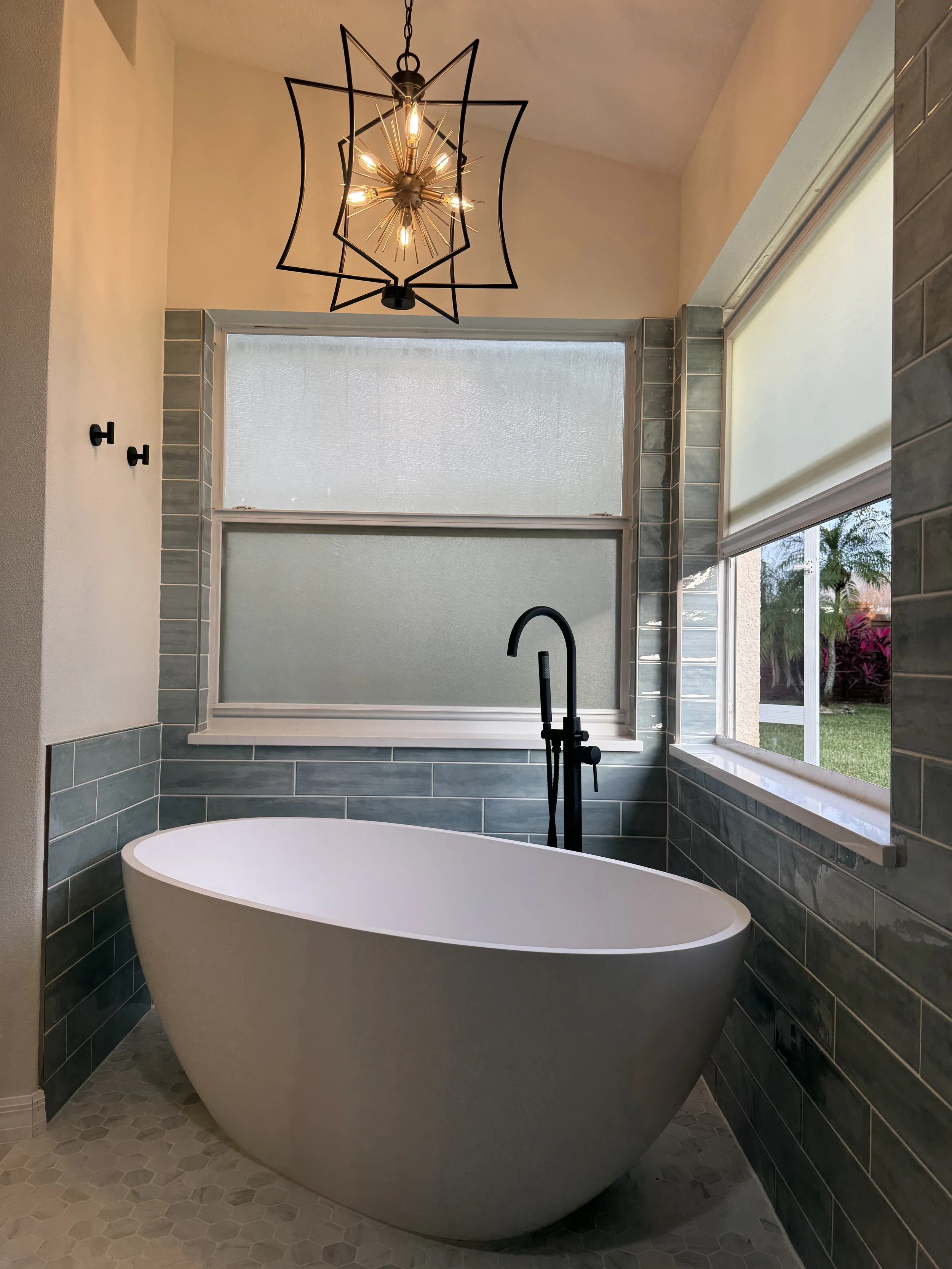 Freestanding soaking bathtub with black floor-mounted faucet, gray subway tile walls, large windows, and decorative pendant lighting in a custom bathroom remodel.