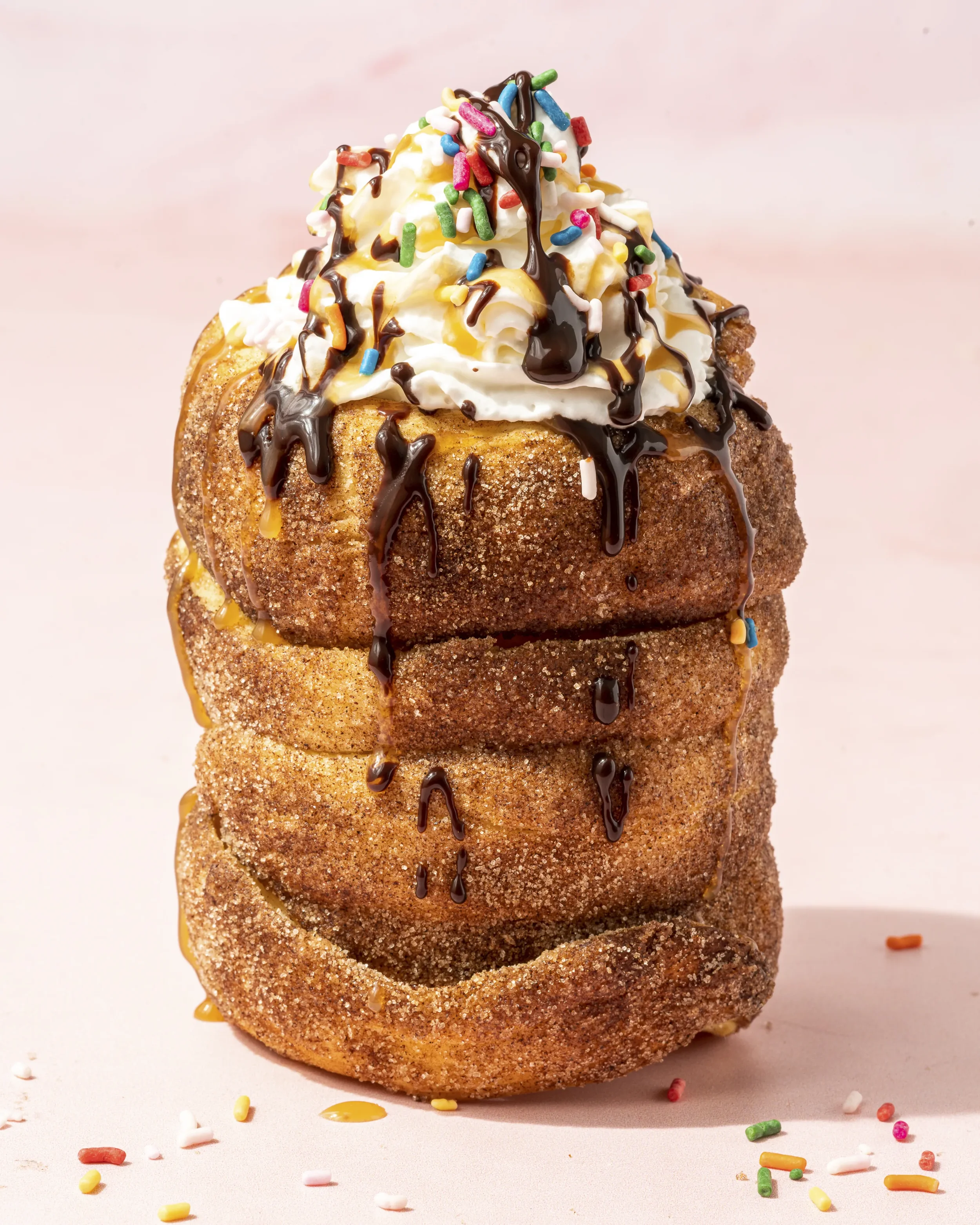 A deep-fried donut topped with whipped cream, colorful sprinkles, caramel sauce, chocolate drizzle, and caramel syrup, with scattered sprinkles around it on a light pink background.