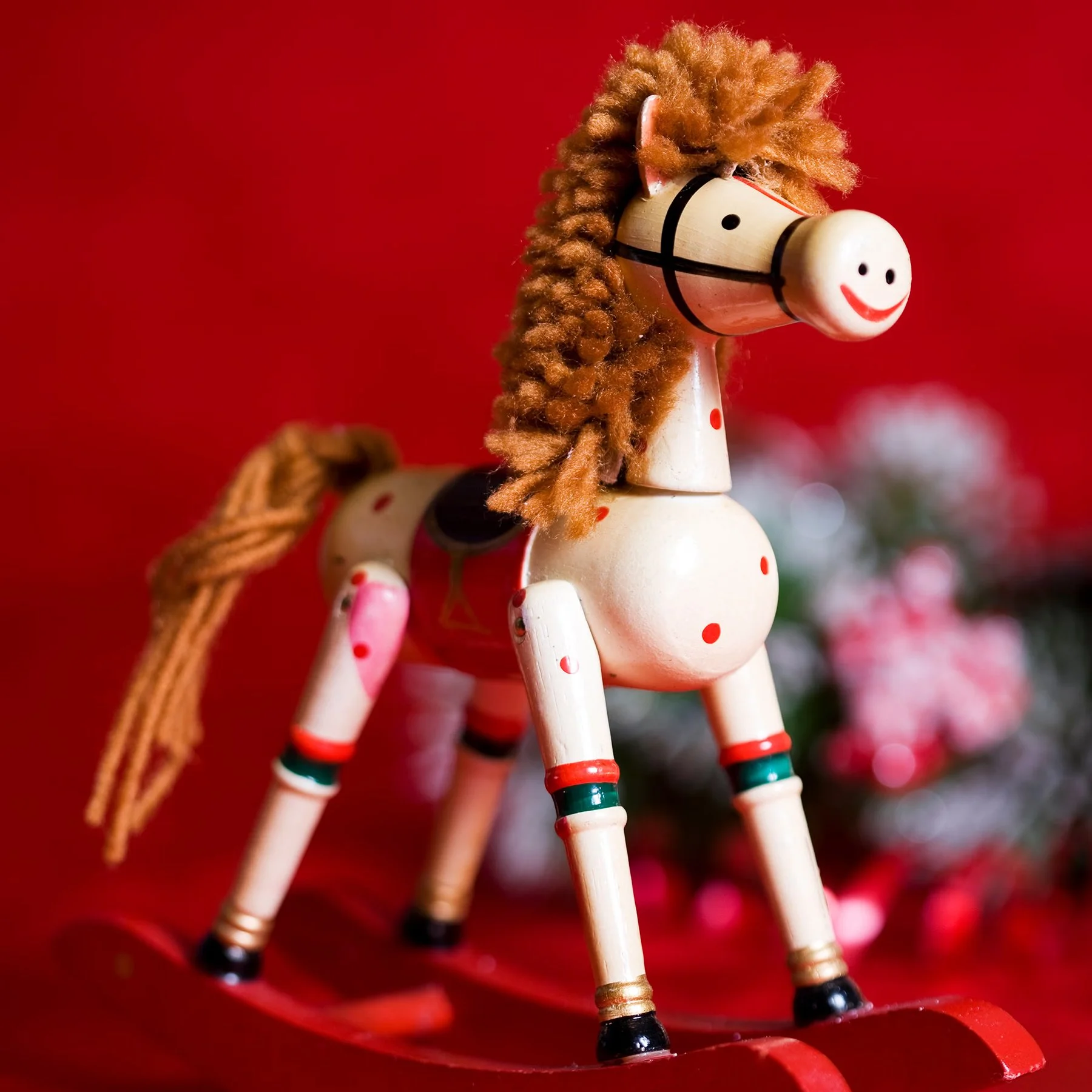Wooden toy horse with a smiling face, brown fuzzy mane, and red and green hooves, on a red background.