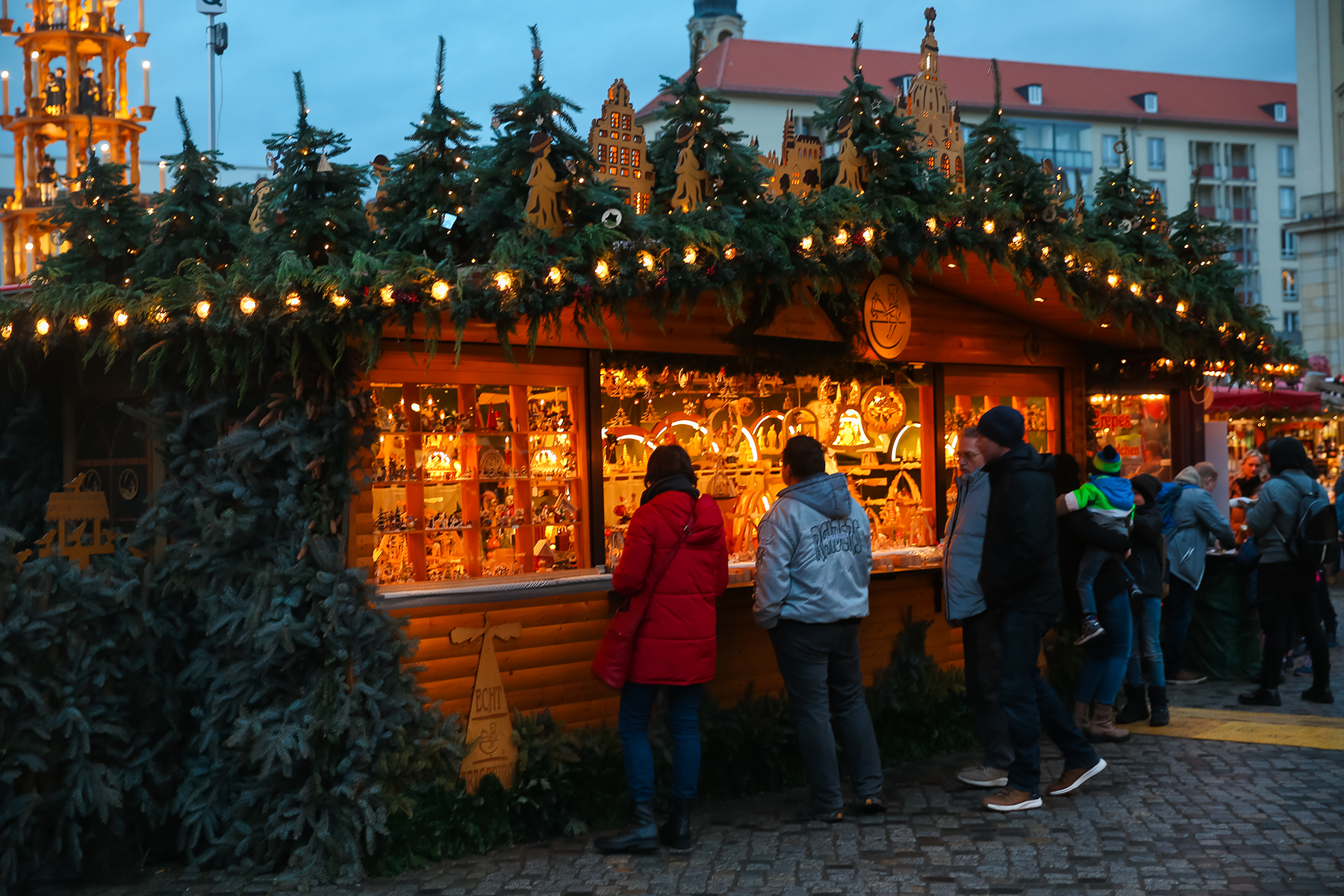 People shopping at a Christmas market stall decorated with holiday lights and greenery, with a large Christmas pyramid in the background.