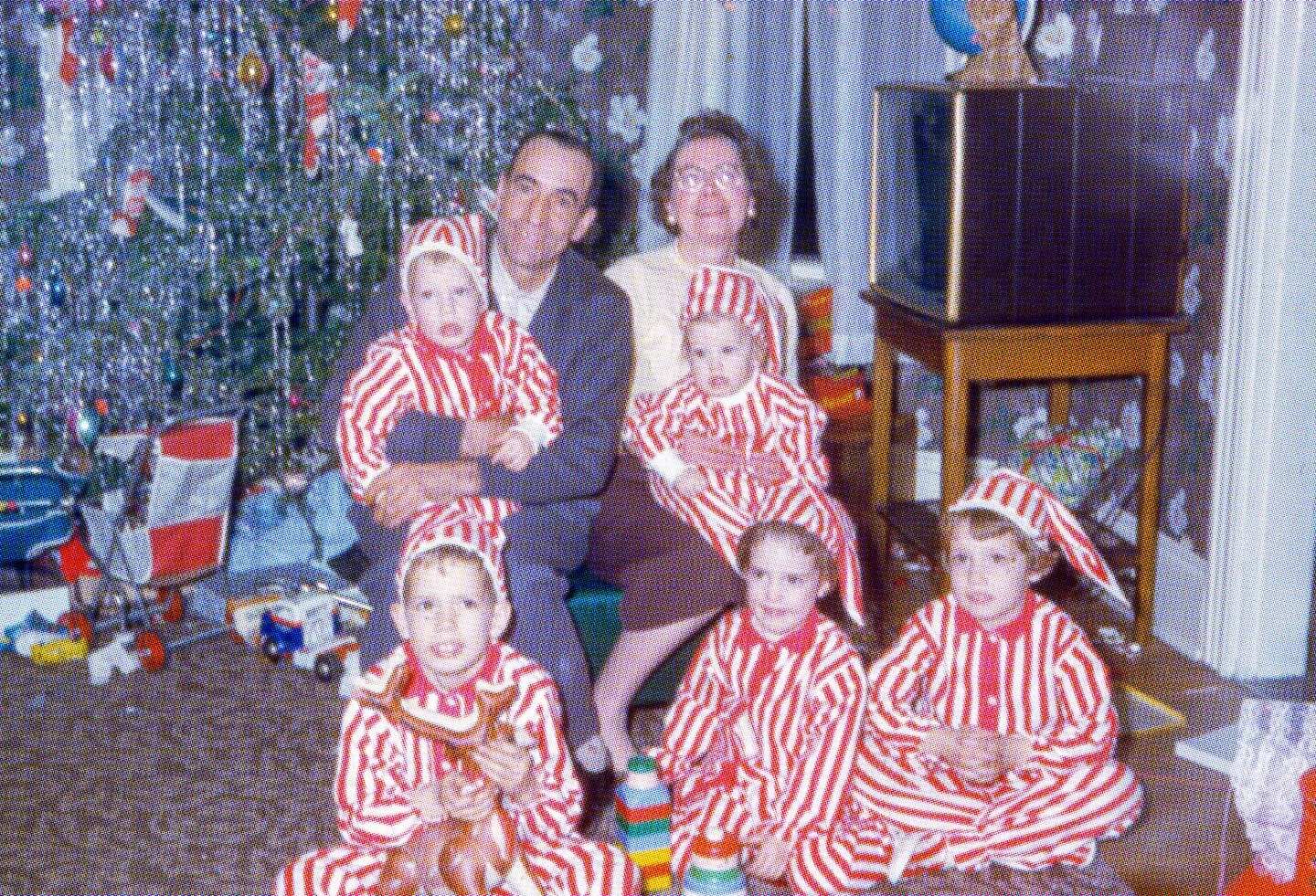 A family dressed in red and white striped Christmas pajamas and hats, gathered in front of a Christmas tree, sitting and standing in a living room.