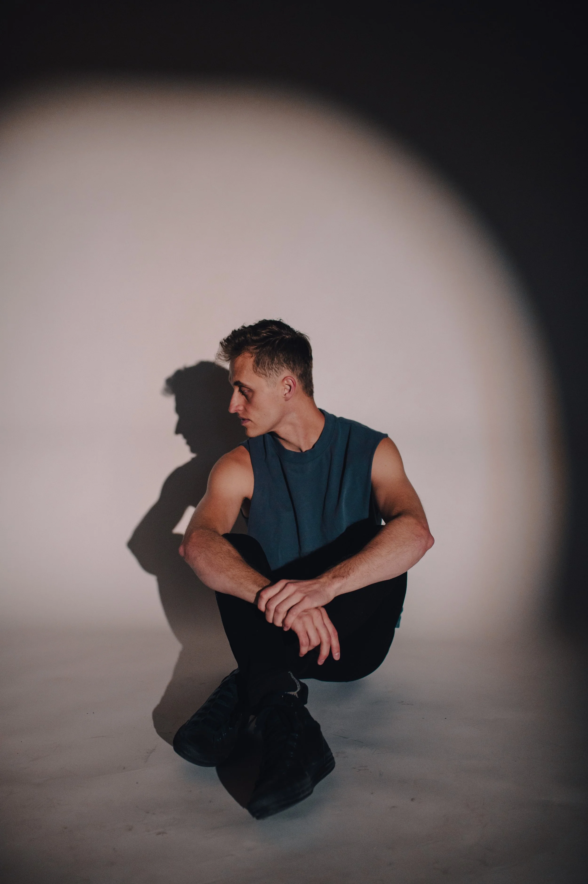 Young man sitting on the floor in a sleeveless shirt, looking to the side against a wall with dramatic shadows.