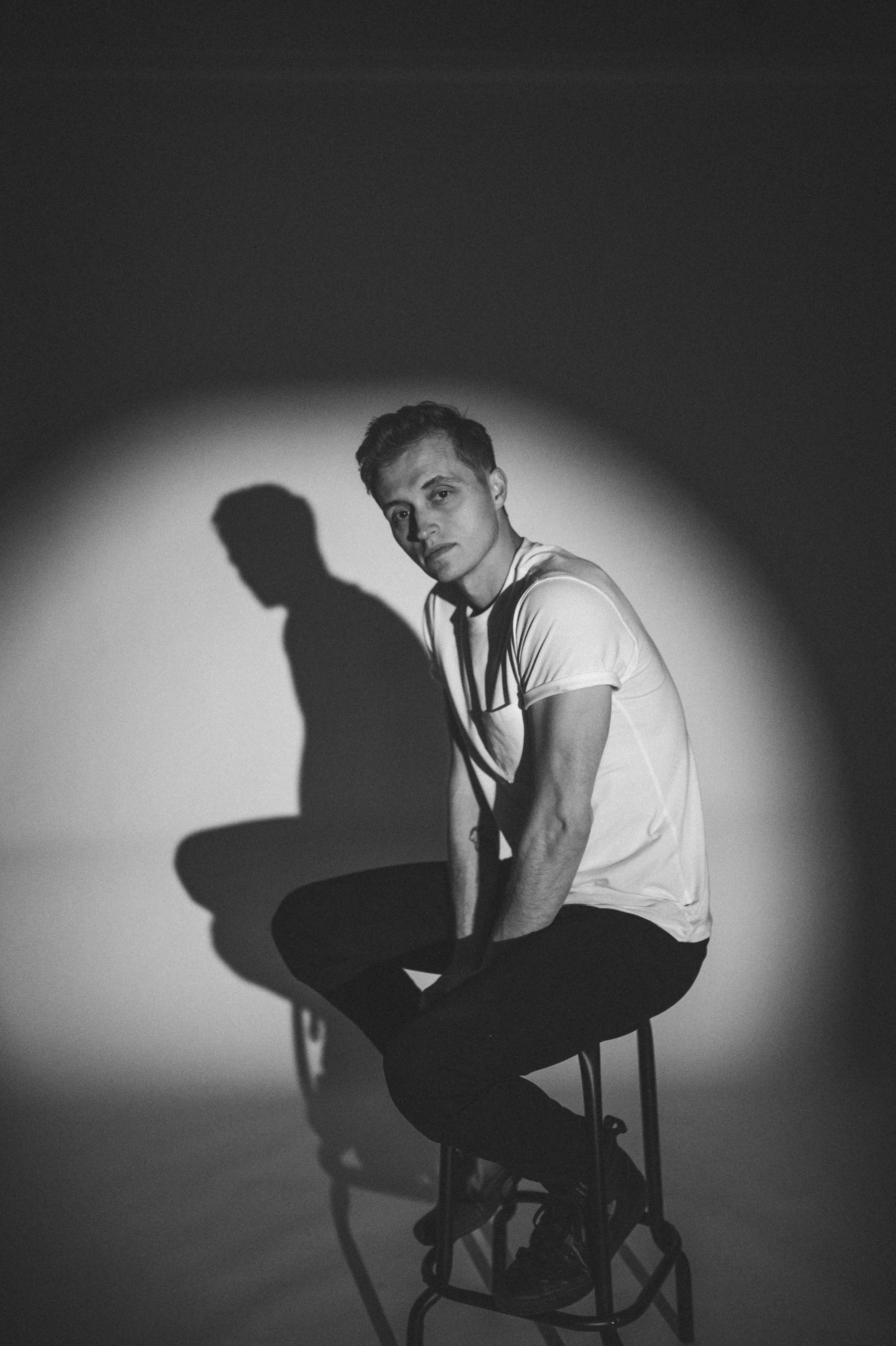 Black-and-white portrait of a young man sitting on a chair against a wall, with dramatic lighting casting his shadow behind him.
