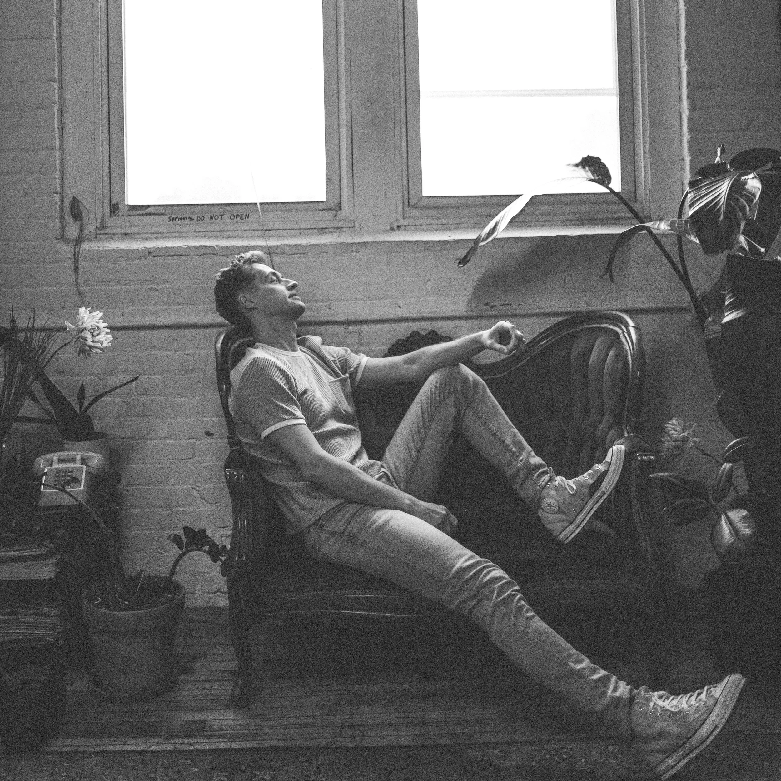 Black-and-white photo of a man reclining in a chair beside a potted plant, with a large bird perched on the chair.