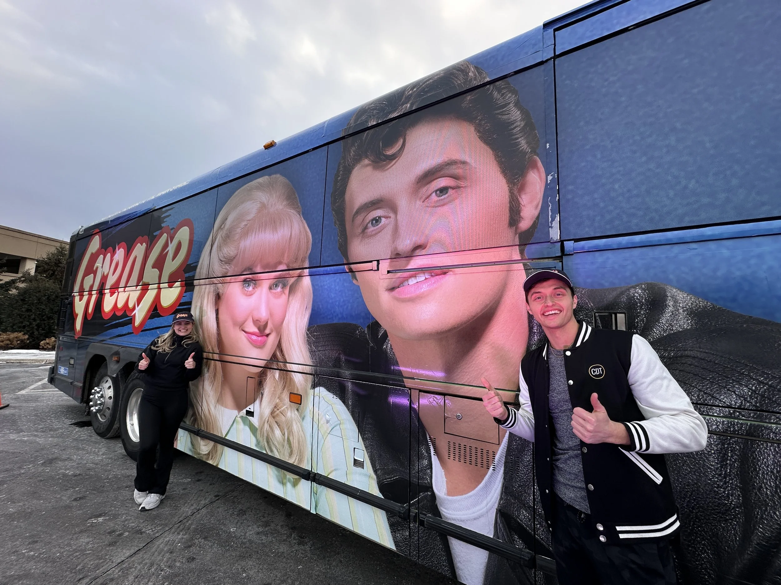 Two people pose beside a tour bus featuring a large Grease movie mural of John Travolta and Olivia Newton-John.