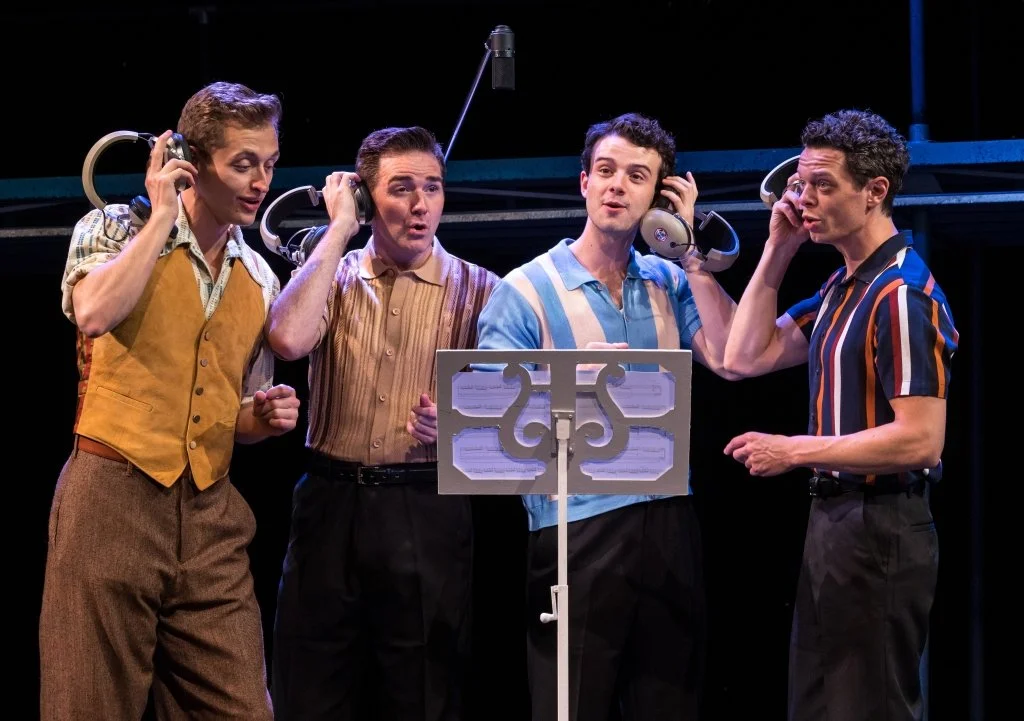 Four male performers wearing headsets sing around a music stand on a theater stage.