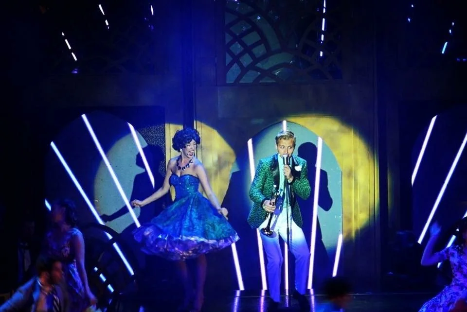 Singers performing on a blue-lit stage, with a woman in a blue dress and a man holding a microphone and trumpet.