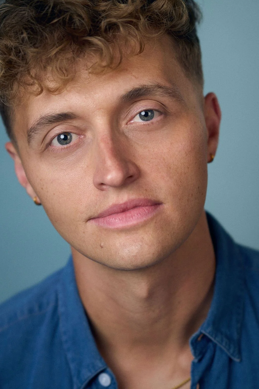 Close-up portrait of a young man with curly brown hair and blue eyes wearing a blue shirt against a teal background.