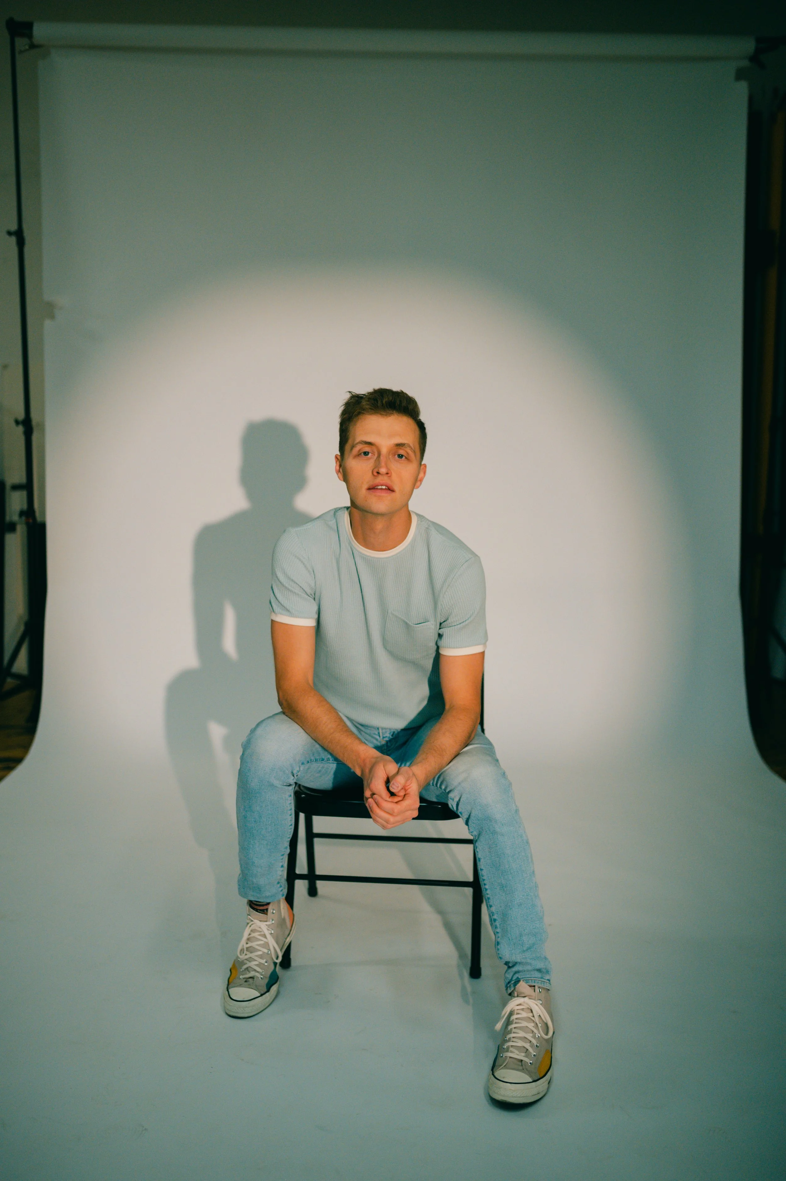 Man sitting on a black chair in a studio, wearing a light blue shirt, jeans, and sneakers.