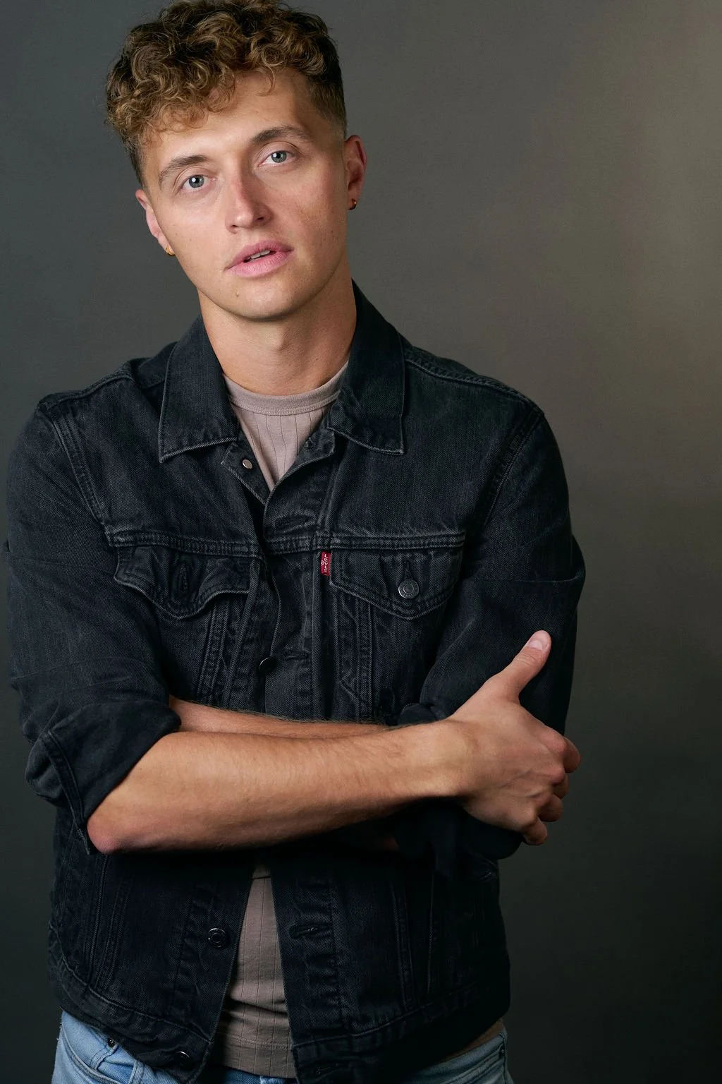 Portrait of a curly-haired man wearing a black denim jacket with his arms crossed against a dark gray background
