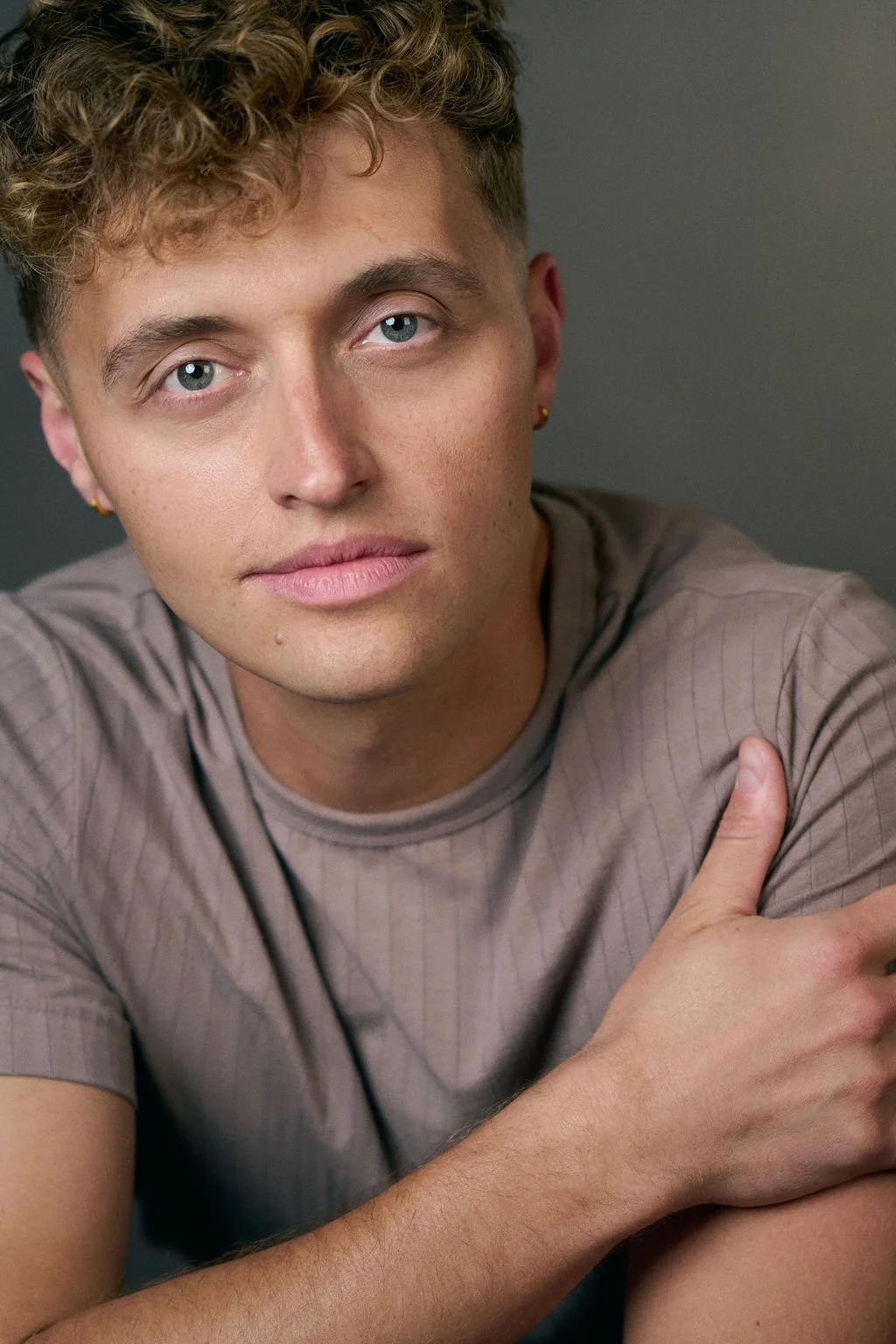 Close-up portrait of a young man with curly hair, blue eyes, earrings, and crossed arms wearing a gray T-shirt