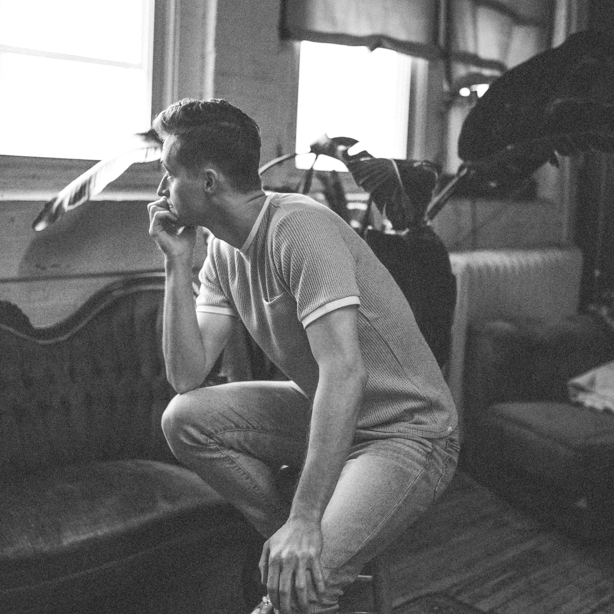Black-and-white photo of a young man sitting on a couch with his hand on his chin, appearing thoughtful.