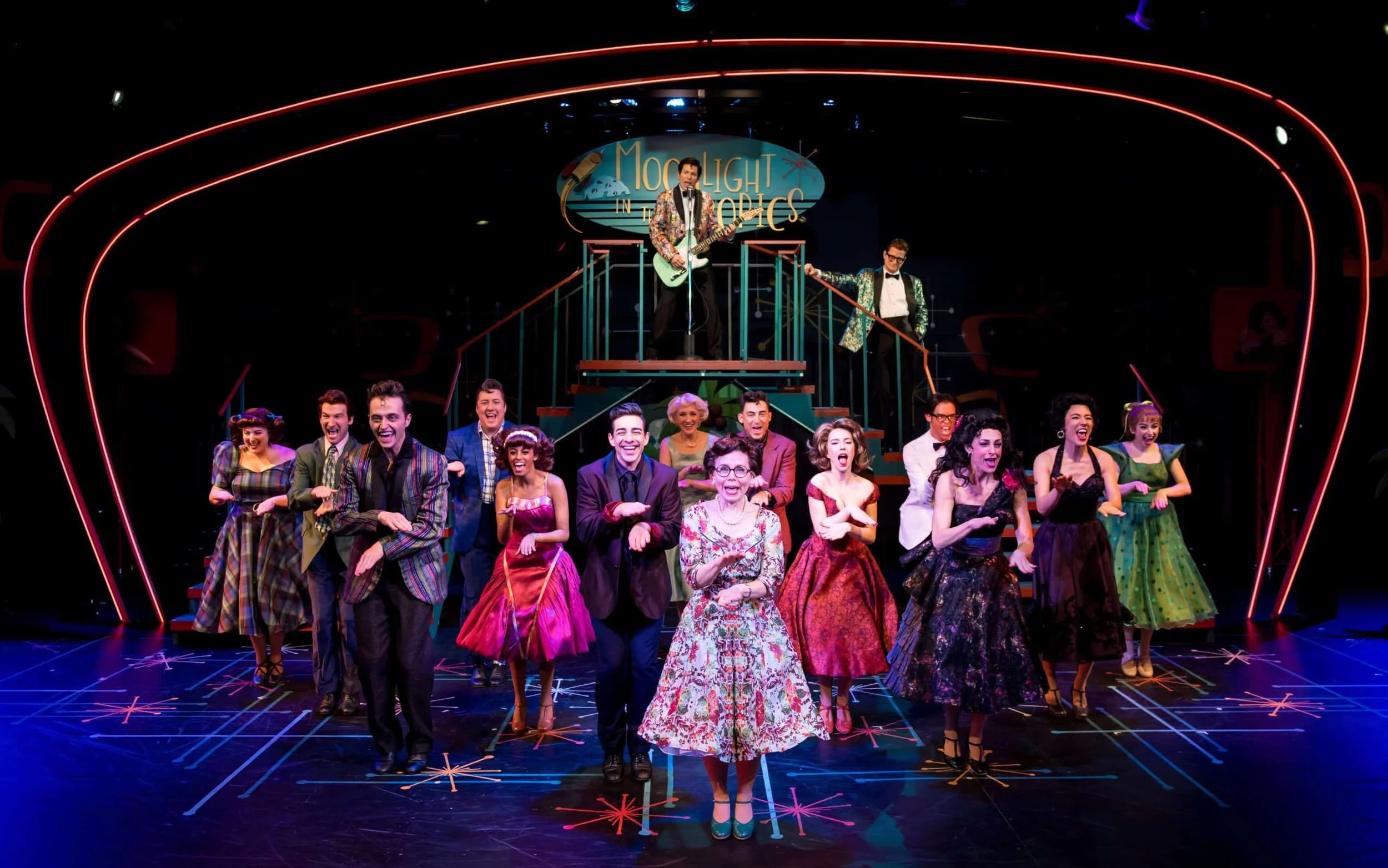 Musical theater cast performing onstage beneath a neon sign reading “Moonlight in the Poconos,” with a guitarist on an elevated platform.