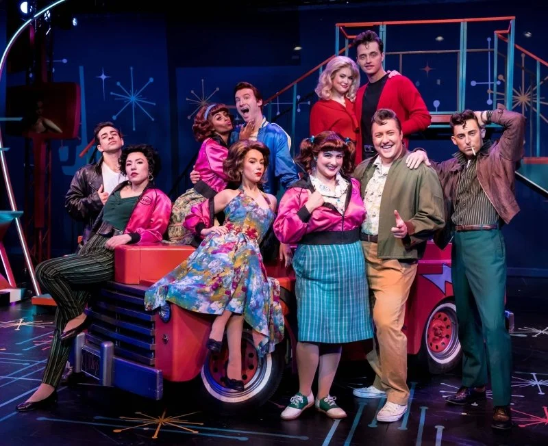 Ensemble cast posing around a colorful vintage vehicle on a retro-themed musical theater stage