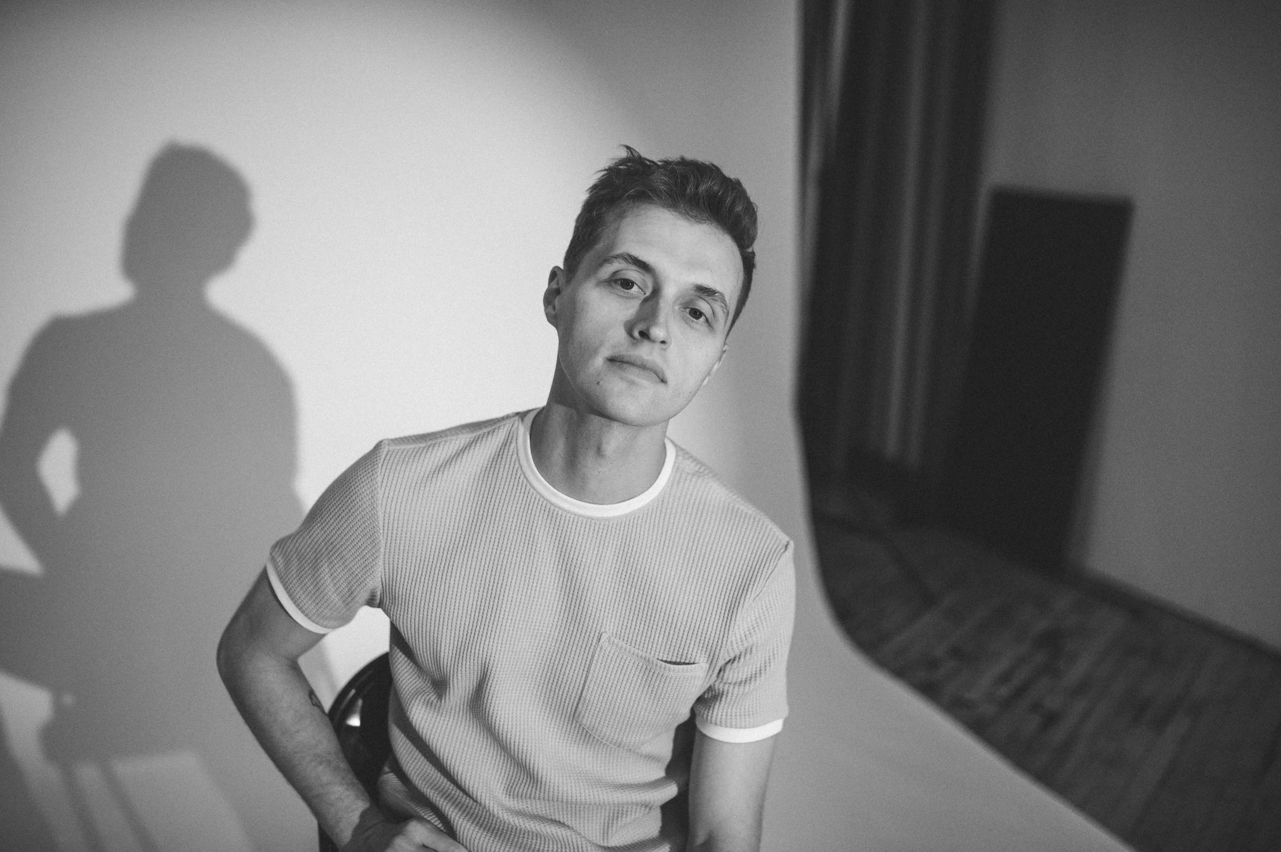 Black-and-white portrait of a man in a ribbed shirt seated indoors, with his shadow cast on the wall.