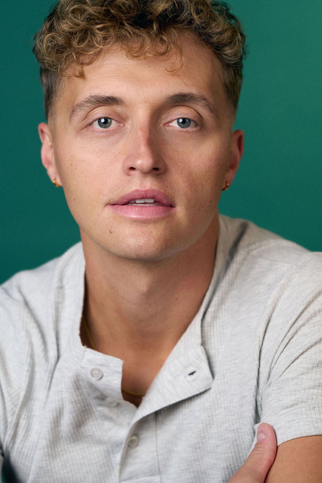 Close-up portrait of a young man with curly brown hair wearing a light gray shirt against a teal background