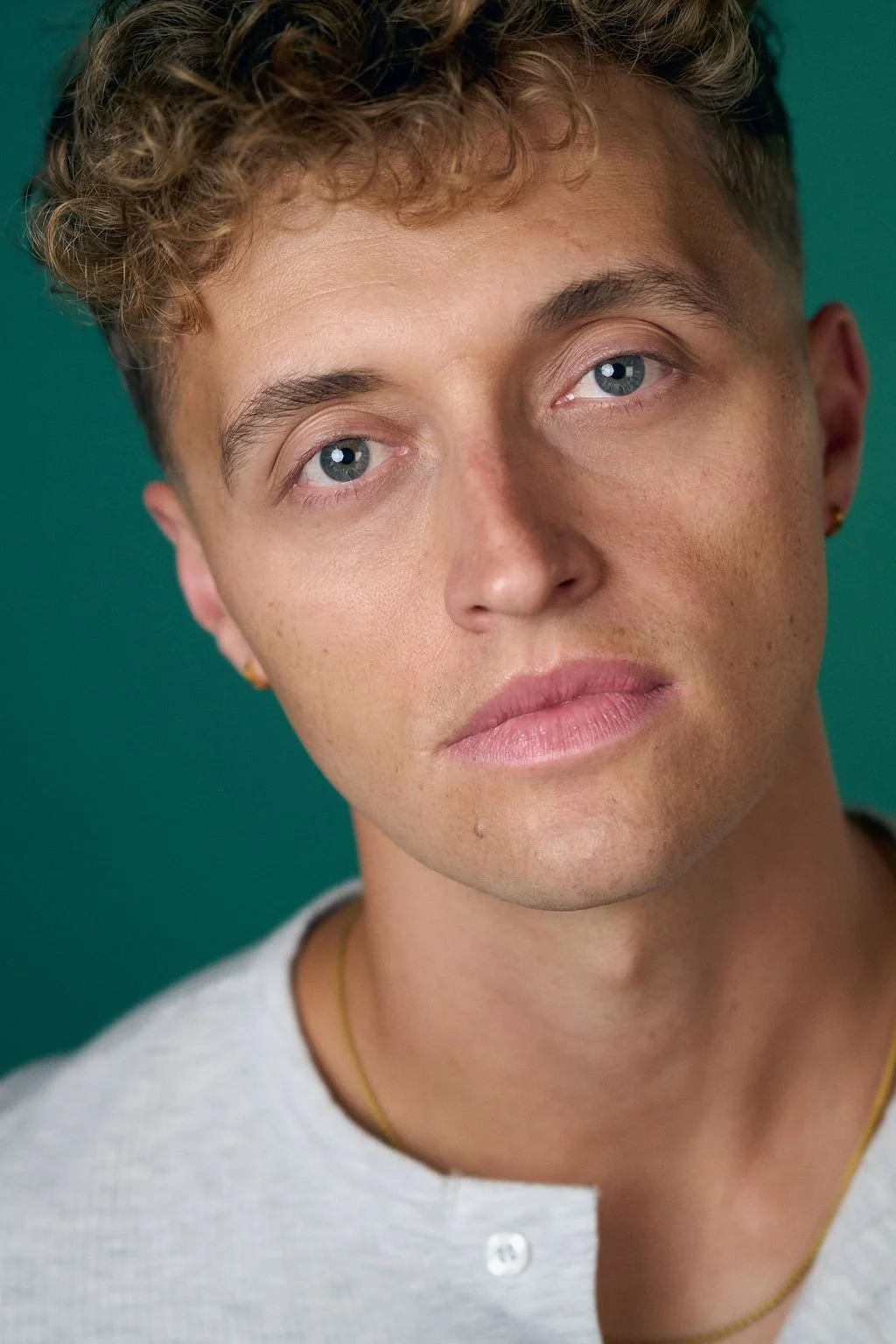 Close-up portrait of a young man with curly hair, blue eyes, gold earrings, and a white shirt against a teal background.