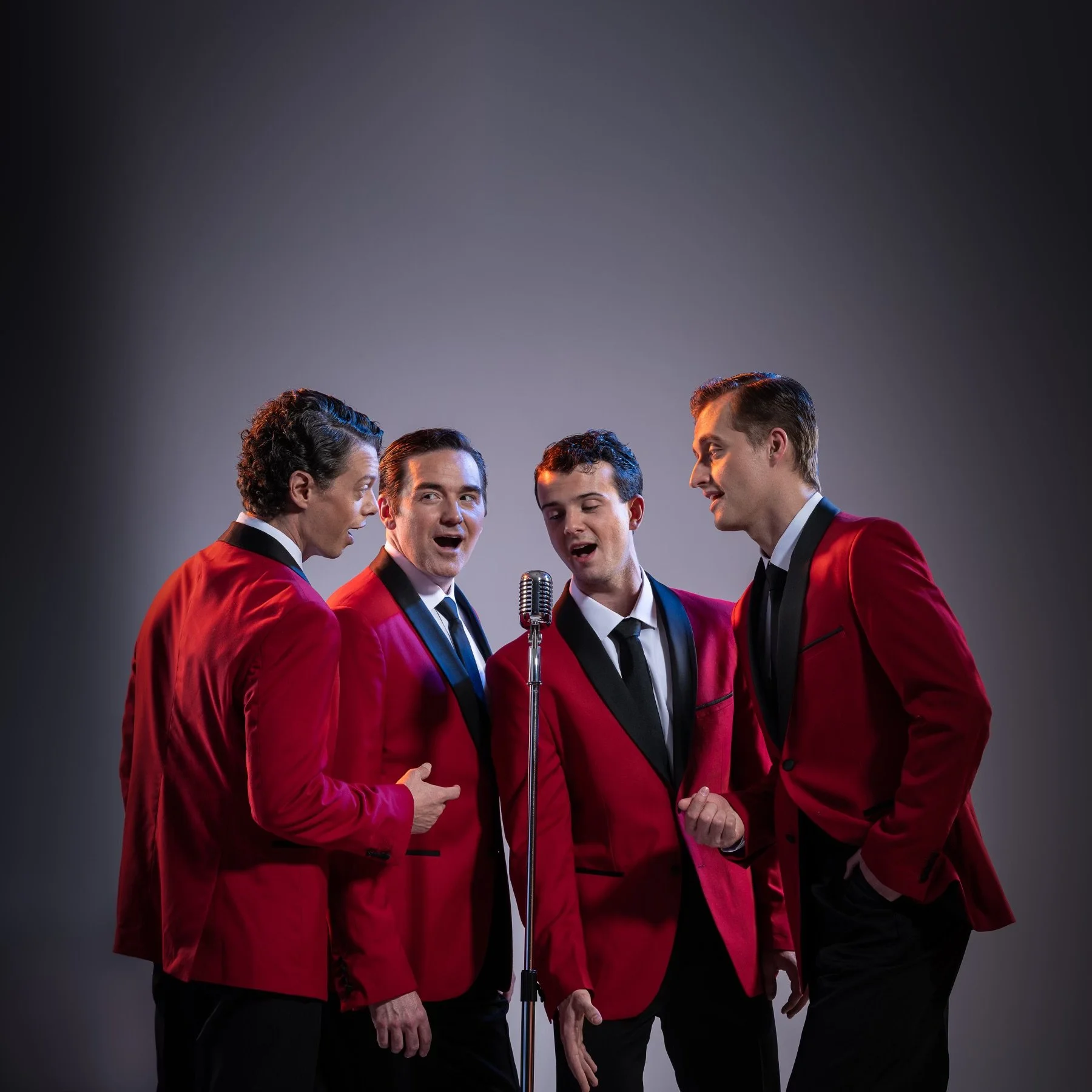 Four men in red tuxedo jackets singing together around a vintage microphone