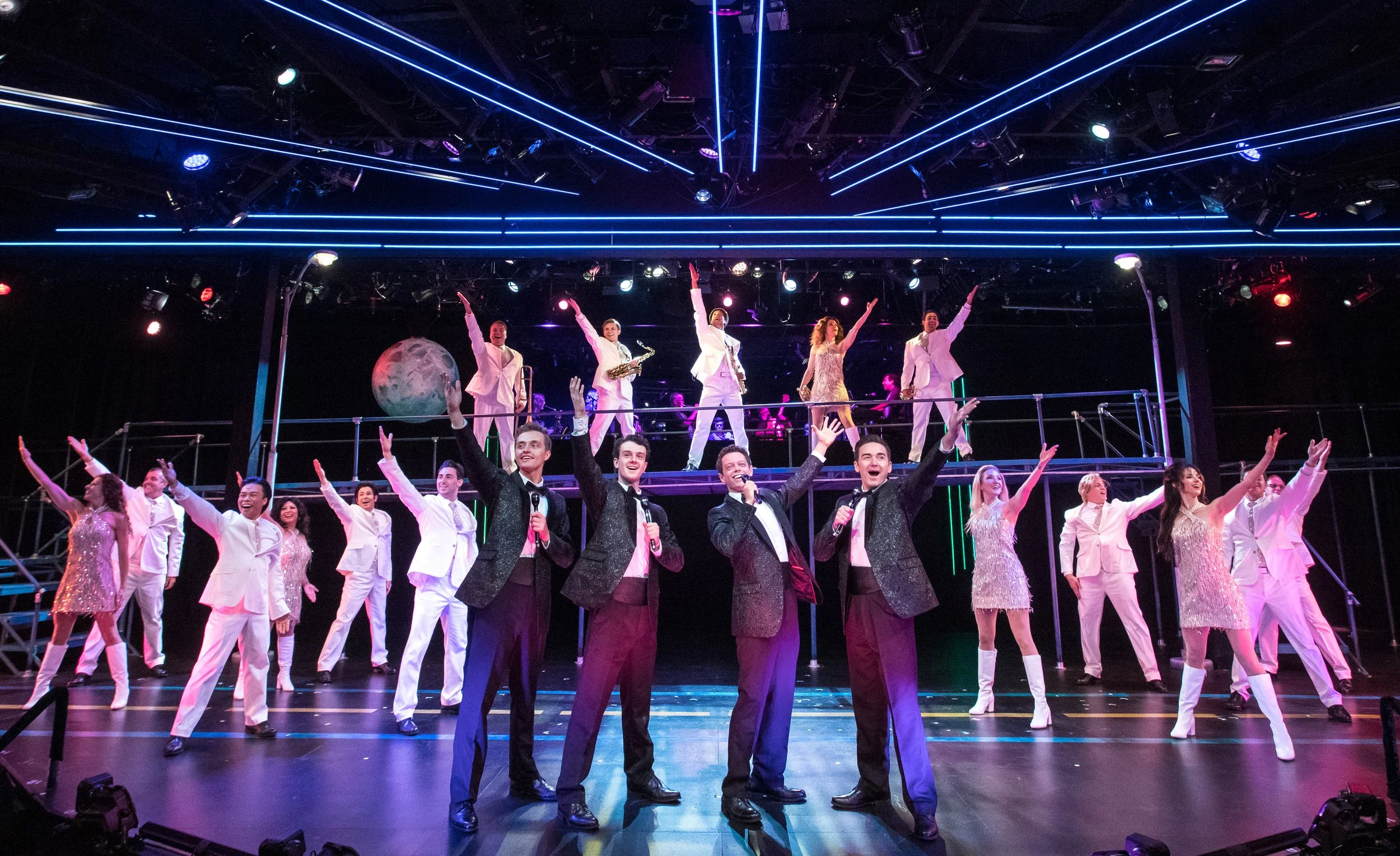Large musical theater ensemble performing under blue and purple stage lights, with singers in tuxedos and dancers in white costumes.