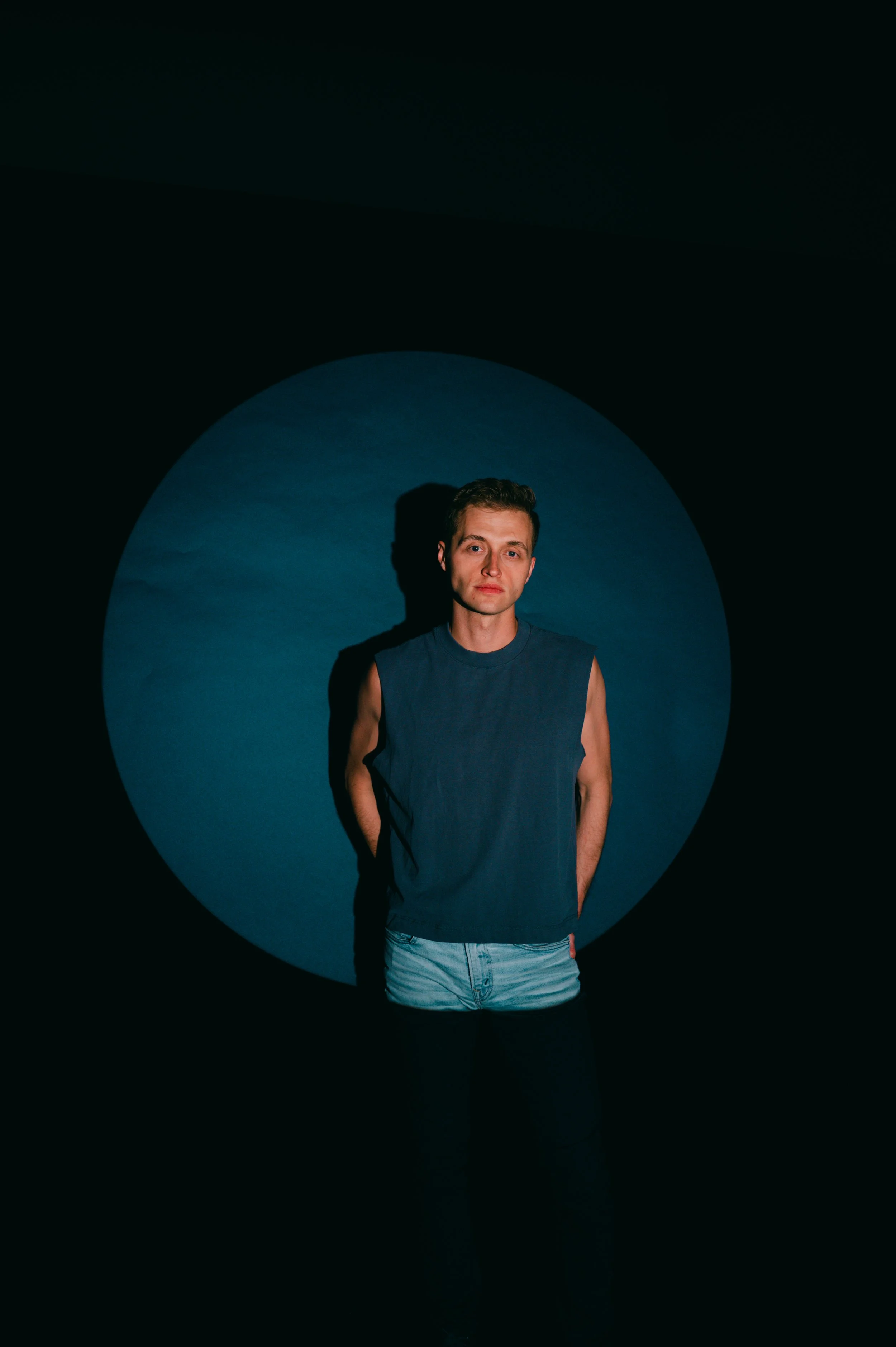 Man in a sleeveless shirt standing against a black background with a large blue circular spotlight and dramatic shadows