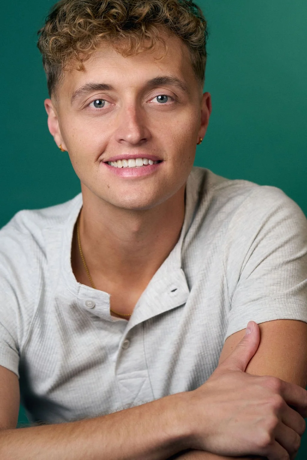 Smiling young man with curly blond hair wearing a light gray shirt against a teal background
