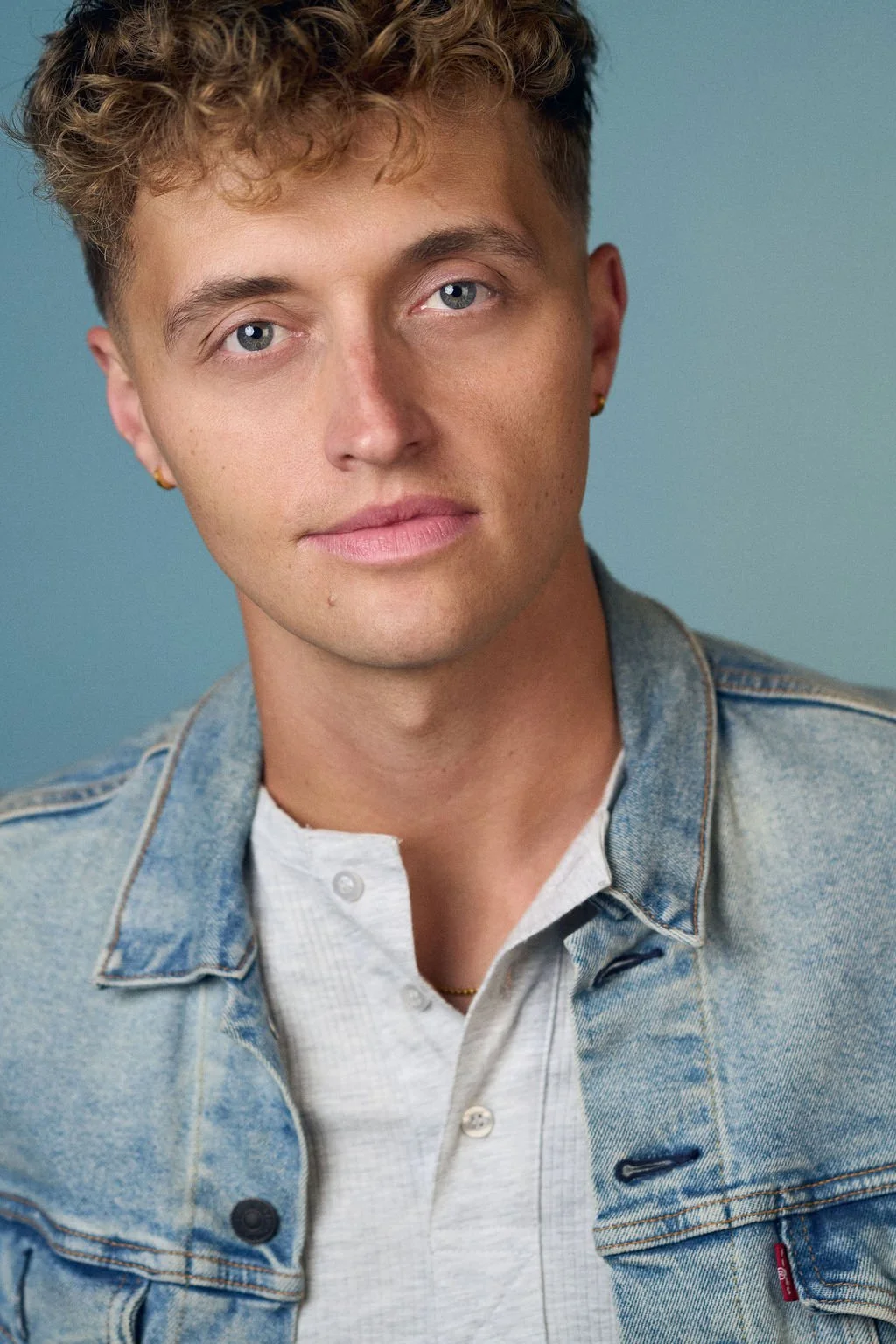 Close-up portrait of a young man with curly brown hair wearing a denim jacket and white shirt against a blue background