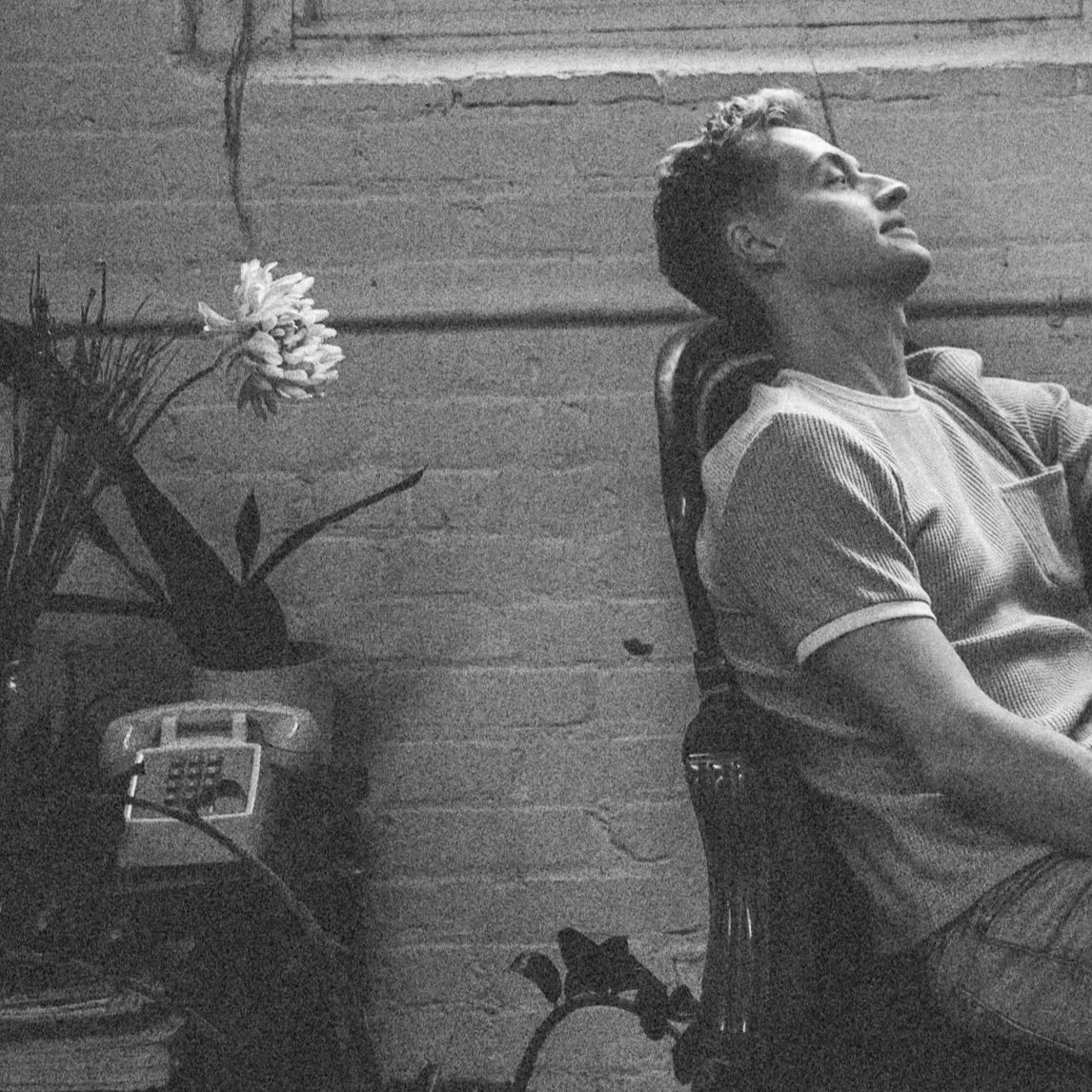 Black-and-white photo of a man reclining beside a potted flowering plant and vintage rotary telephone