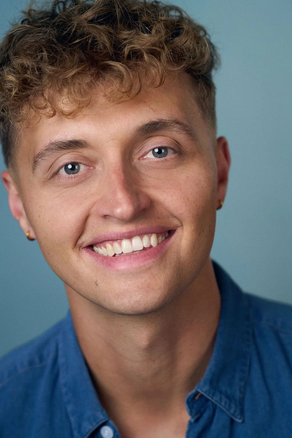 Close-up portrait of a smiling young man with curly brown hair, blue eyes, earrings, and a blue shirt against a teal background.