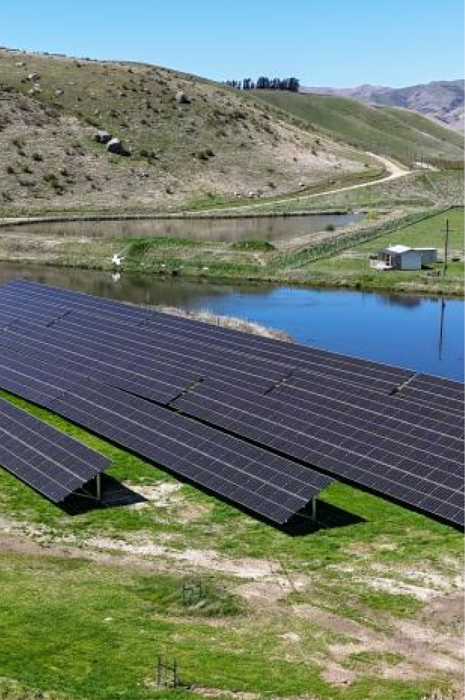 Solar panels installed on a grassy field near a body of water in a hilly landscape.