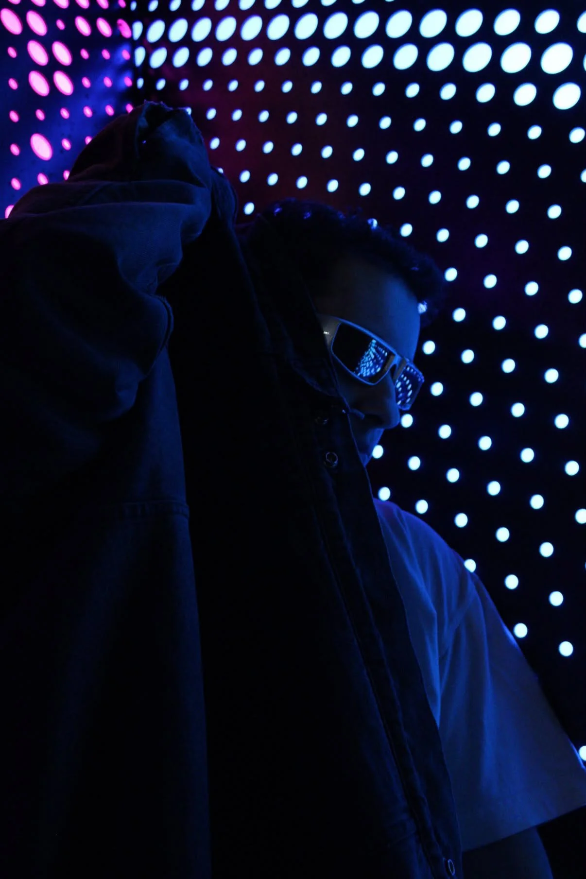 A person wearing glasses and a dark jacket is standing in front of a backdrop of blue and pink LED lights that create a dotted pattern.