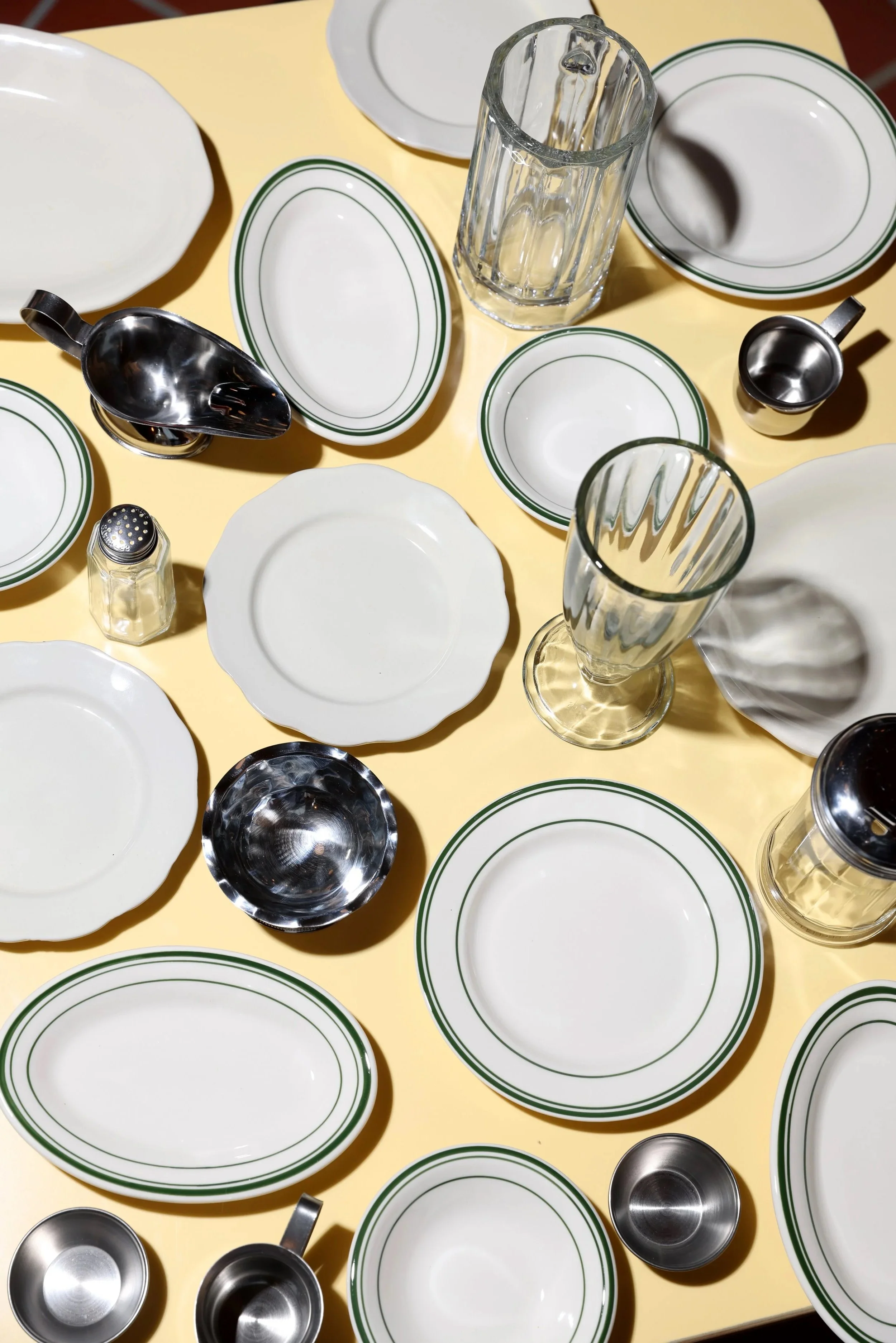 Multiple empty white plates with green borders, clear glass vases, small silver cups, and a salt shaker on a yellow table.