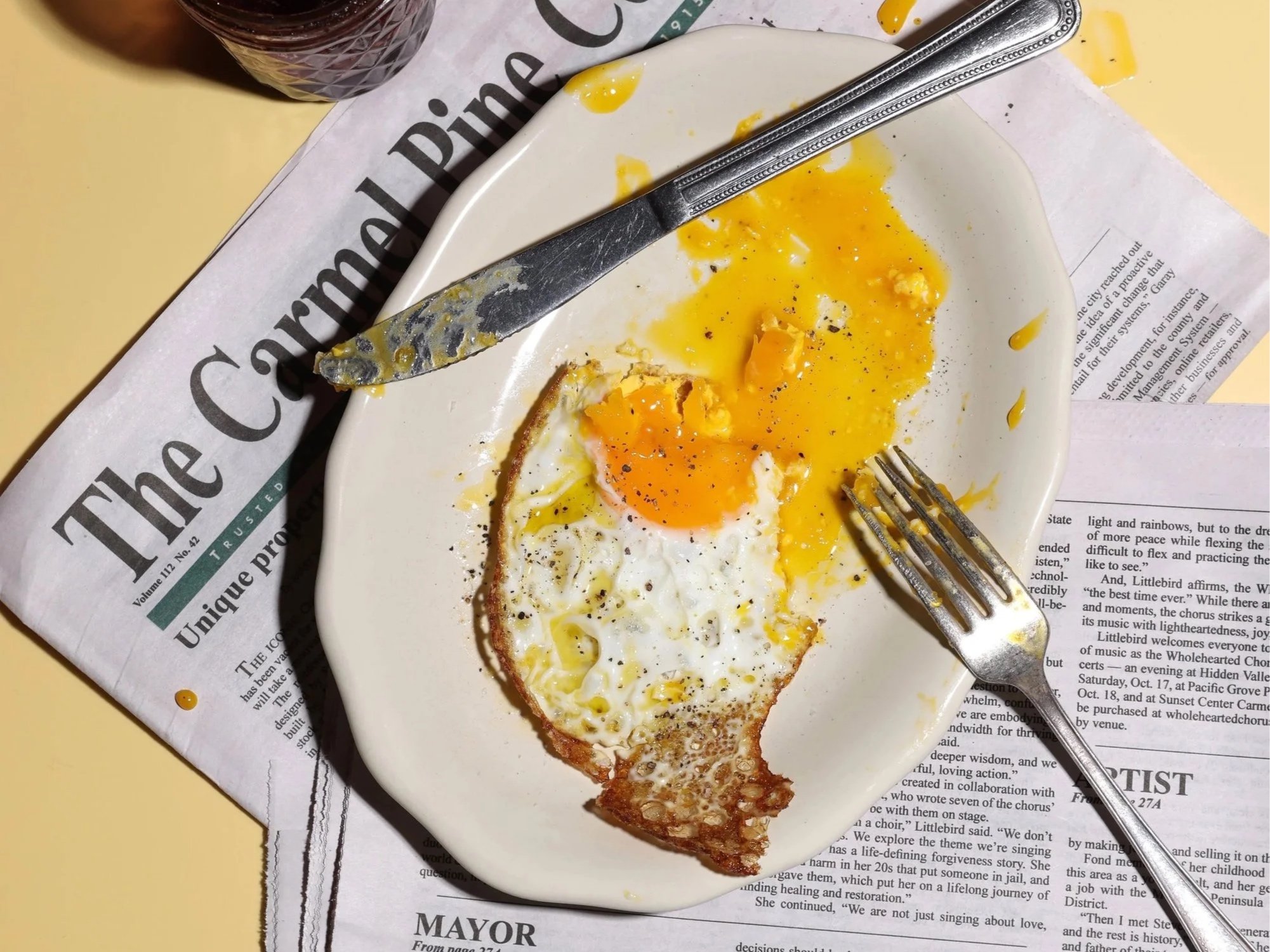 A breakfast plate with a fried egg and a slice of toast, partially eaten. The plate is on a newspaper with a fork and a knife resting on the plate, and a glass of dark beverage in the background.
