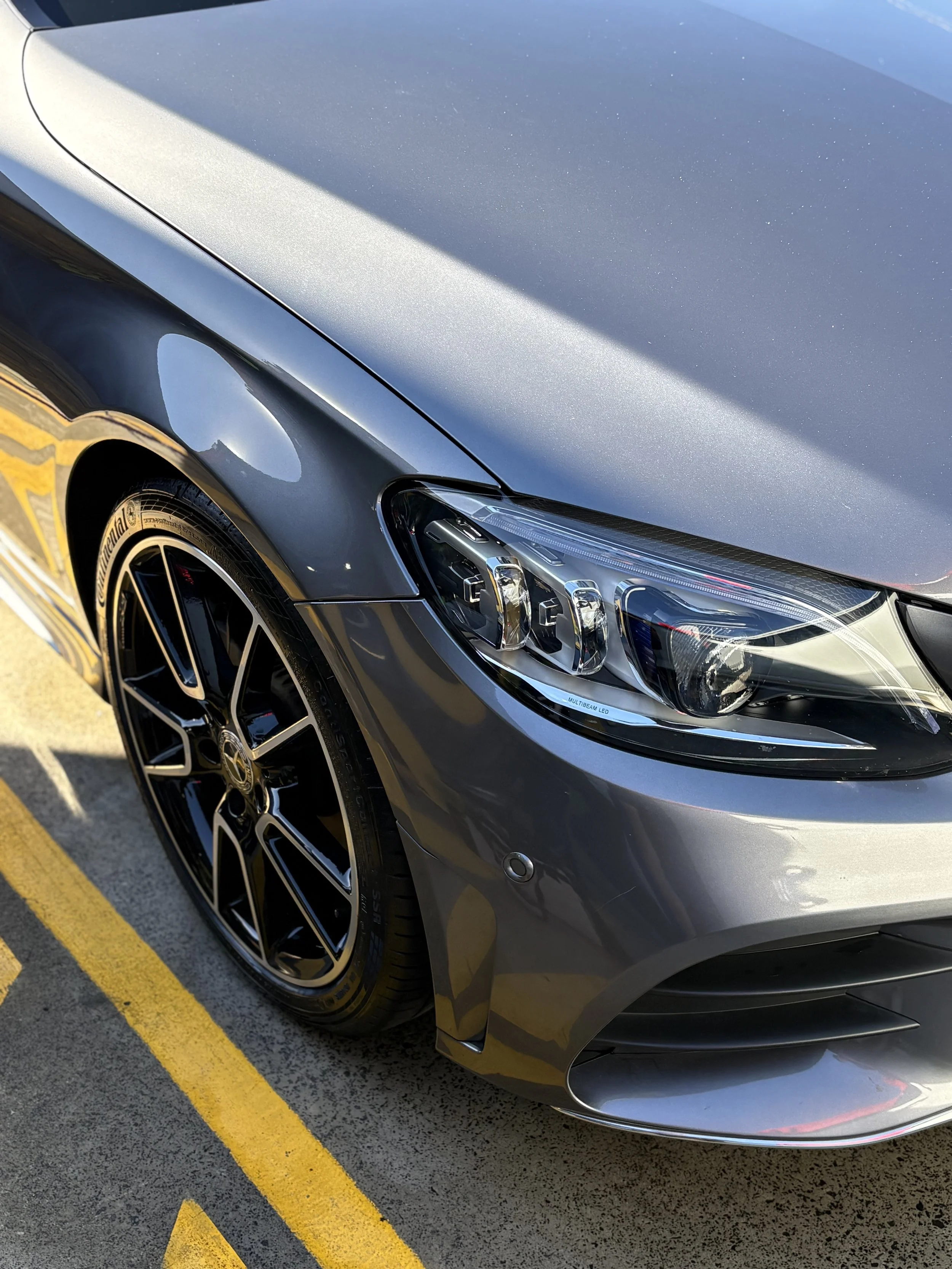 Close-up of a gray modern car parked in a parking lot with yellow lines, showing the front right side including the headlight, wheel, and part of the hood.