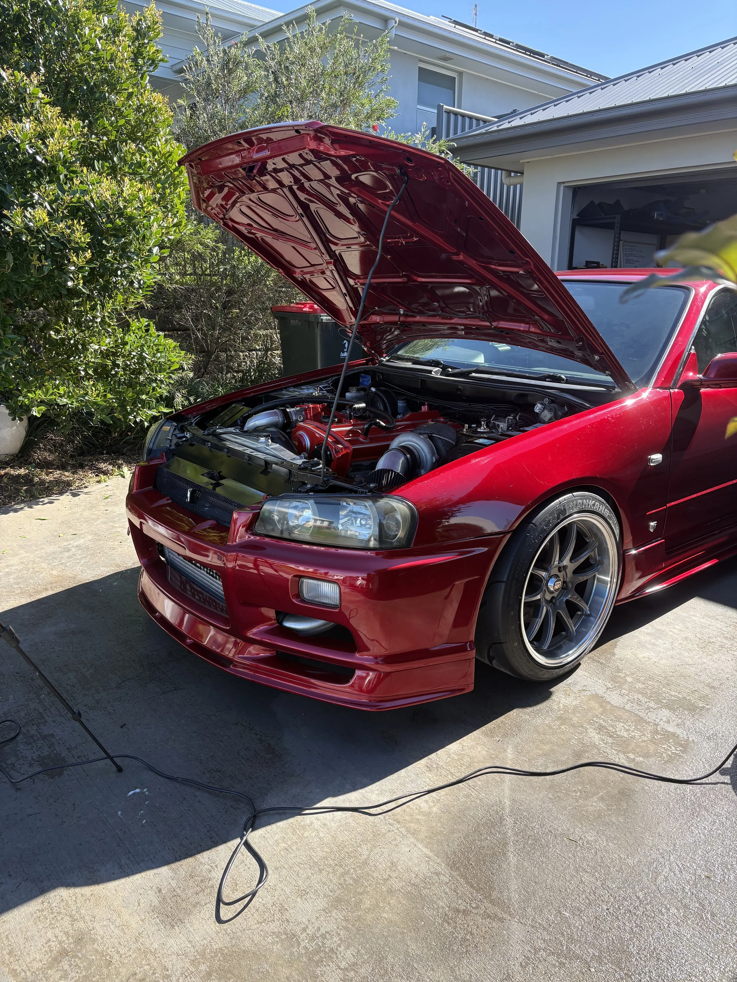 A red sports car with its hood open, exposing the engine, parked outside a house on a sunny day.
