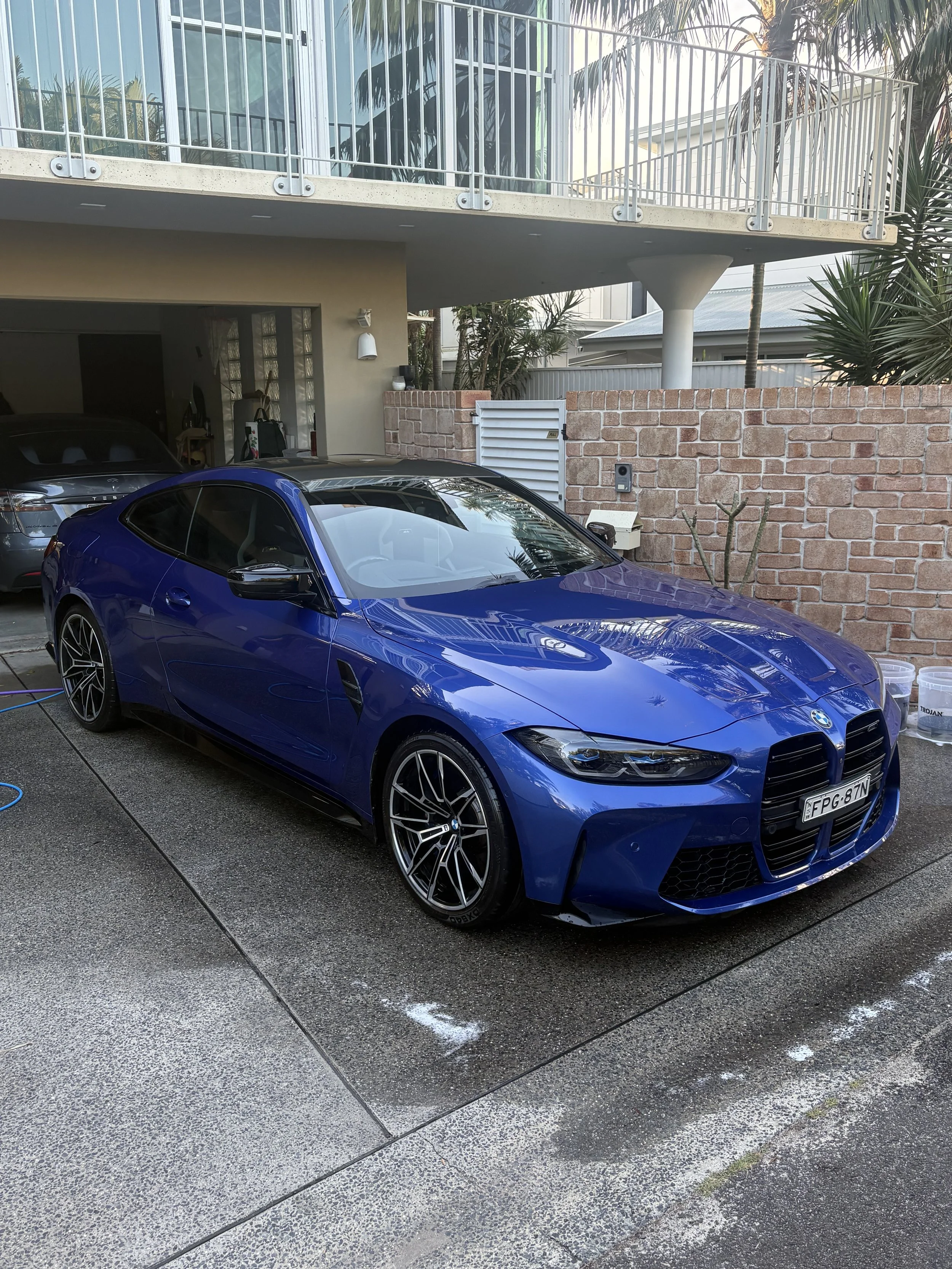 A blue BMW sports car parked in a driveway next to a house with a brick wall, with another gray car parked behind it. The area appears to be a residential home with a balcony and tropical plants.